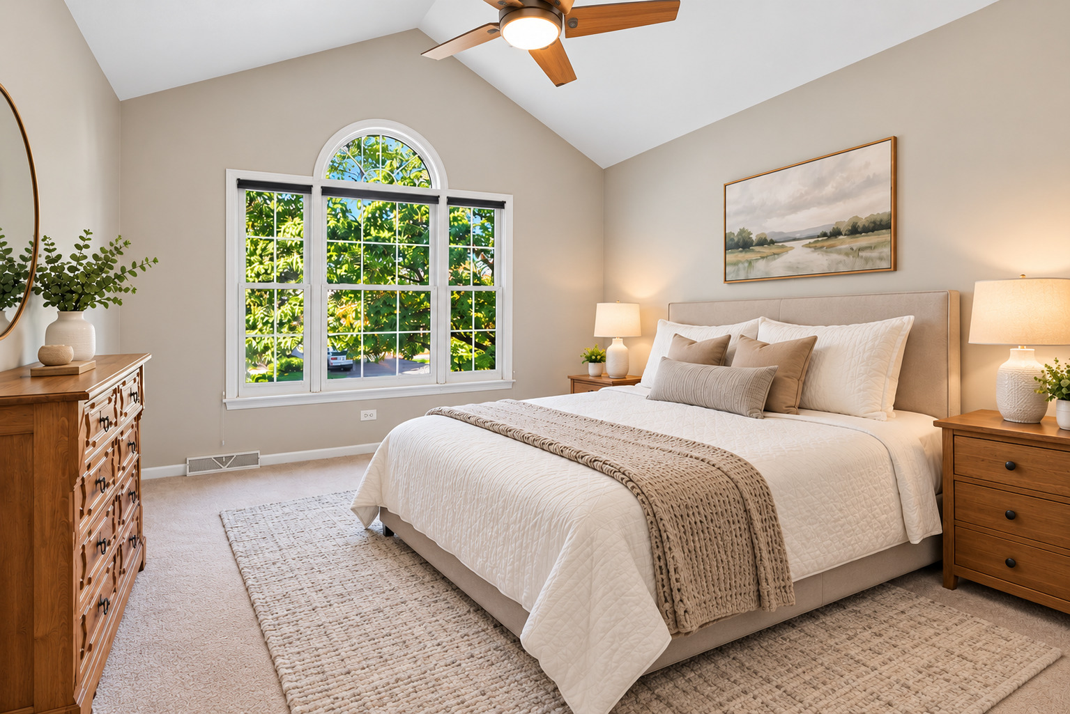 Staged bedroom with a king-size bed, vaulted ceiling, and arched window overlooking leafy trees.