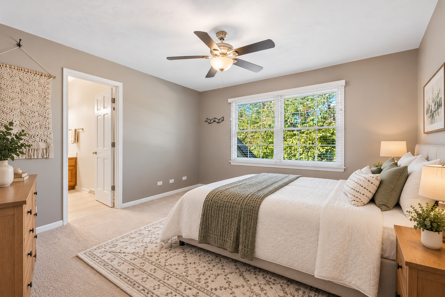 Staged bedroom with a white bed, ceiling fan, and large window overlooking leafy trees.