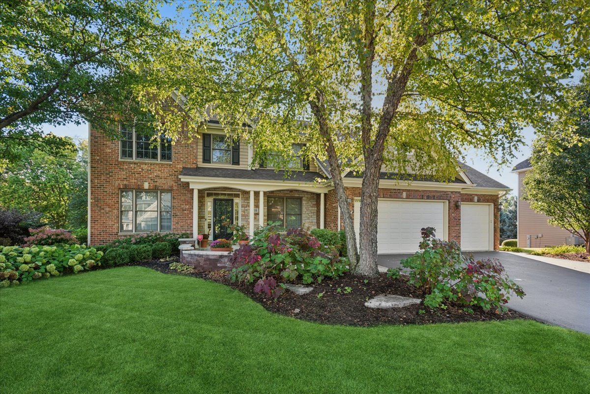 Two-story brick home with an attached garage, mature trees, and landscaped front yard.