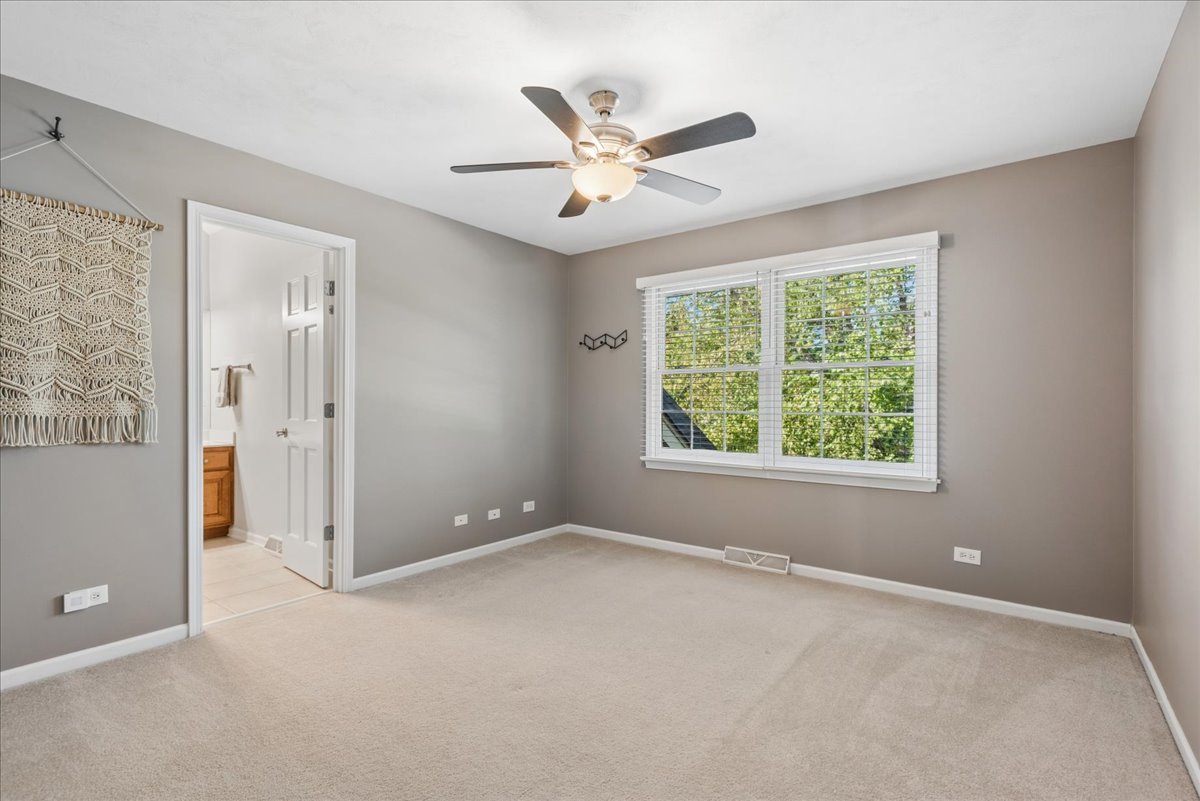 Empty bedroom with beige carpet, ceiling fan, and a large window overlooking leafy trees.