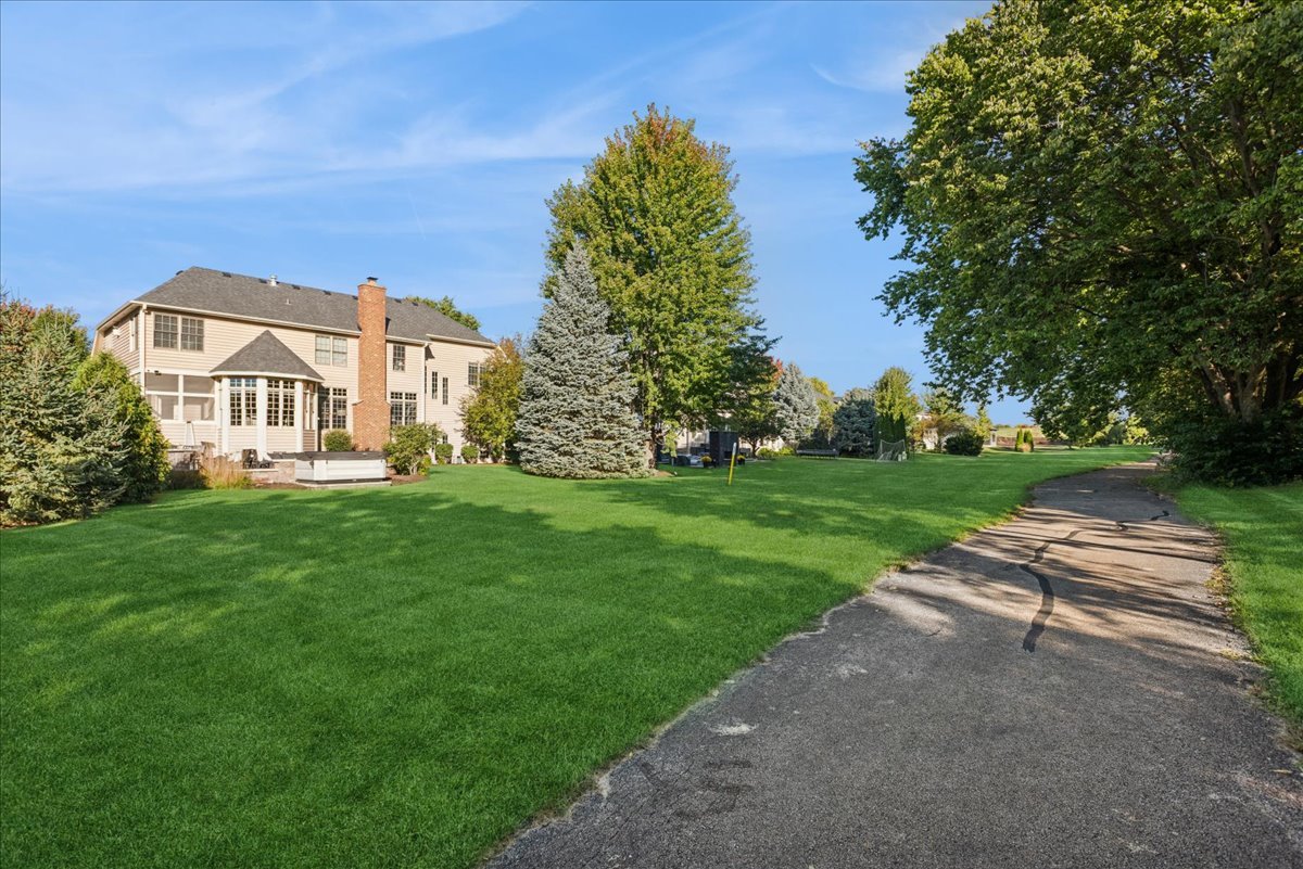 Two-story beige house with a brick chimney beyond a broad lawn and curving paved drive.