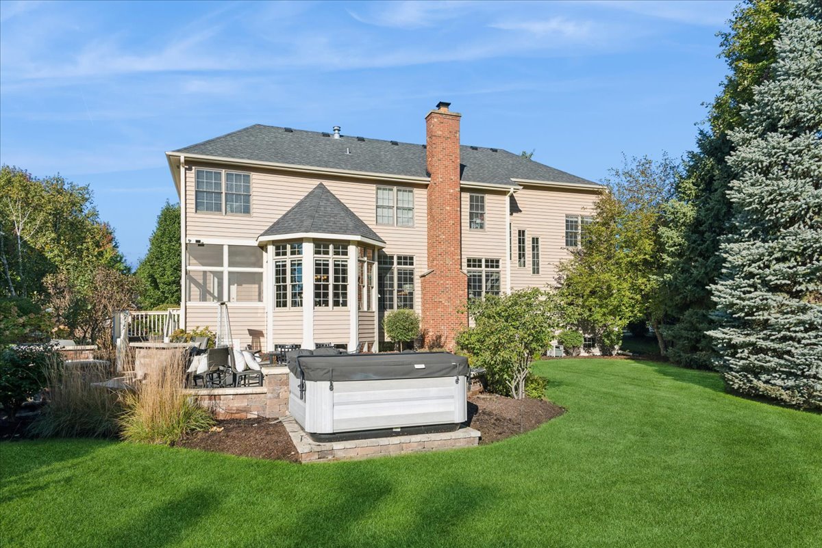 Home’s rear exterior with a brick chimney, bay-windowed sunroom, patio hot tub, and spacious lawn.