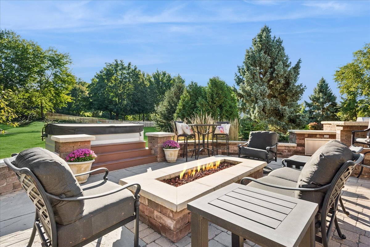 Backyard patio with a stone fire pit, cushioned chairs, raised hot tub, and built-in fireplace.
