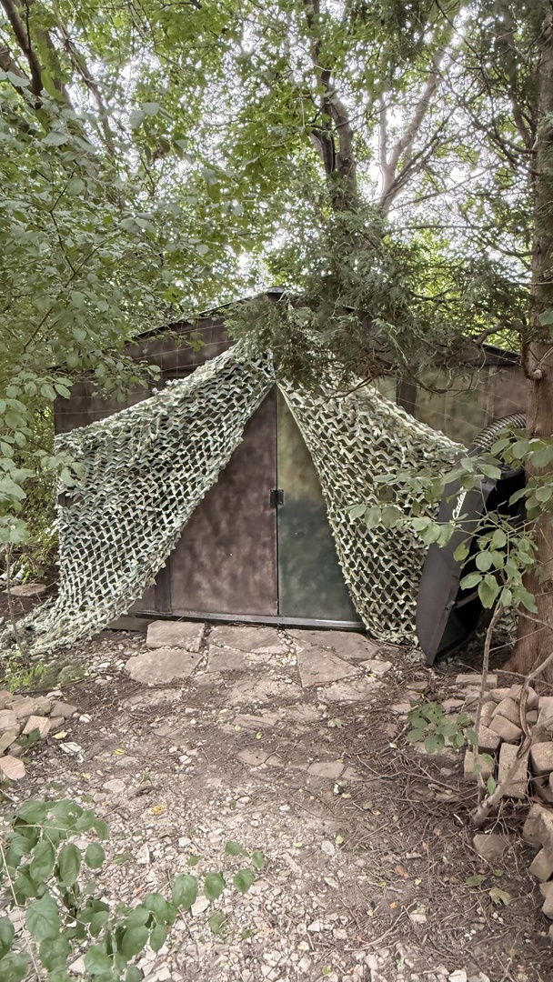 Small outbuilding draped in camouflage netting beneath trees, beside an irregular stone path.