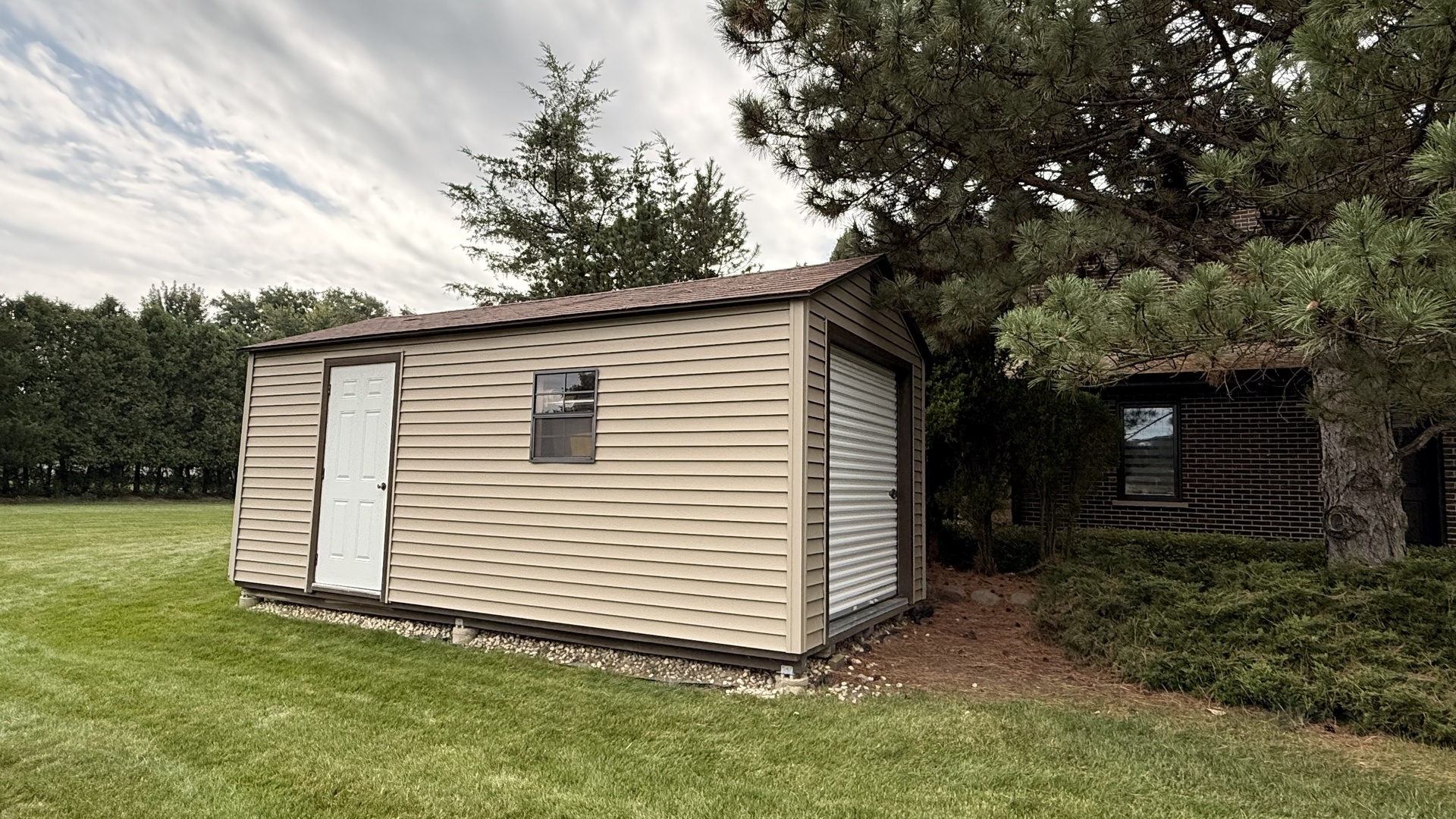 Tan detached shed with a white entry door and roll-up garage door beside a brick house.