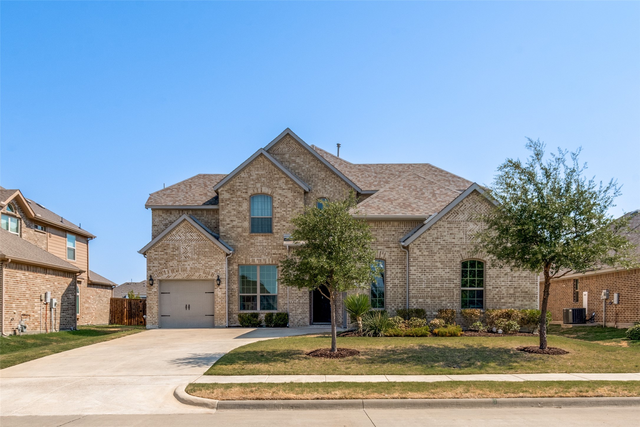 Two-story tan brick home with a double garage, gabled roof, and landscaped front yard.