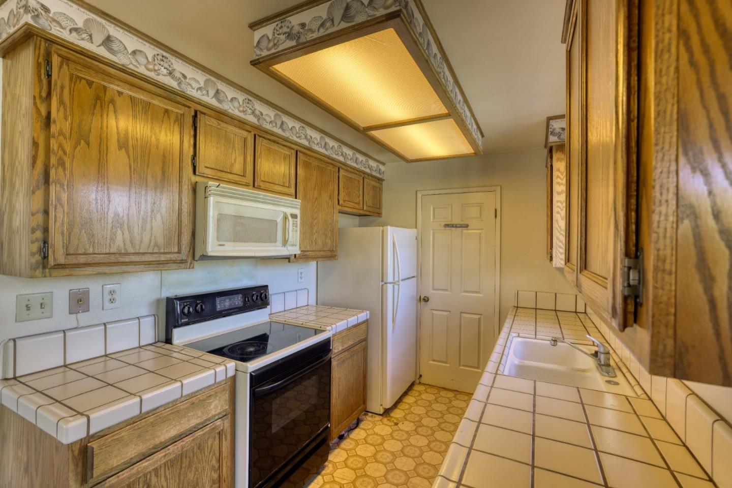 Galley kitchen with oak cabinets, tiled counters, white appliances, and a patterned seashell wall border.