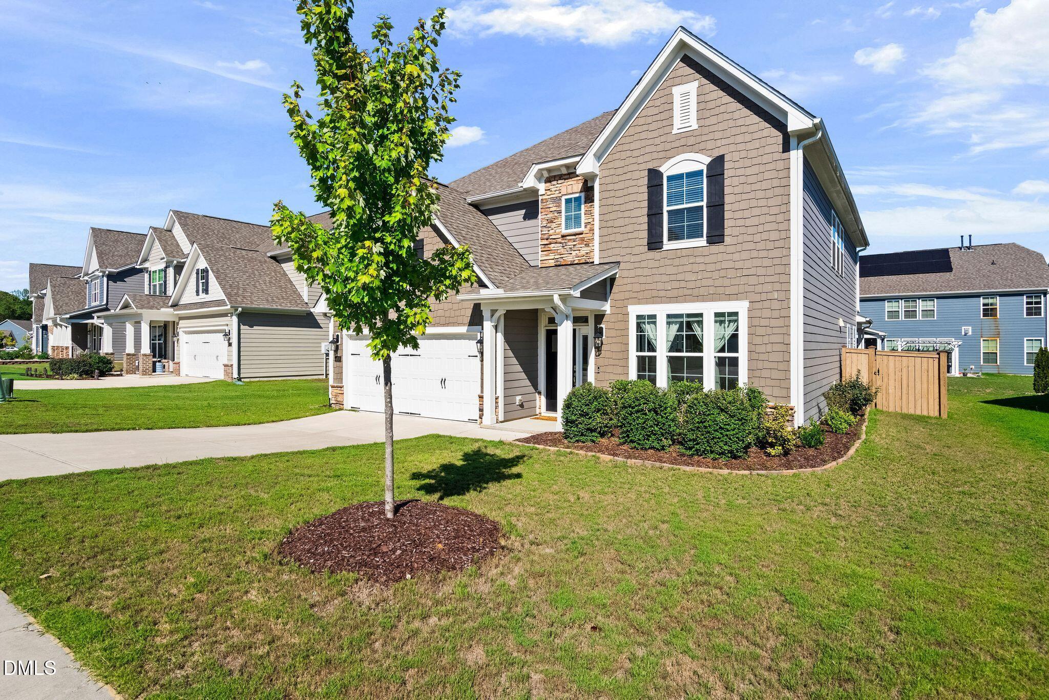 Two-story taupe home with stone accents, white double garage, and a young tree in the front lawn.