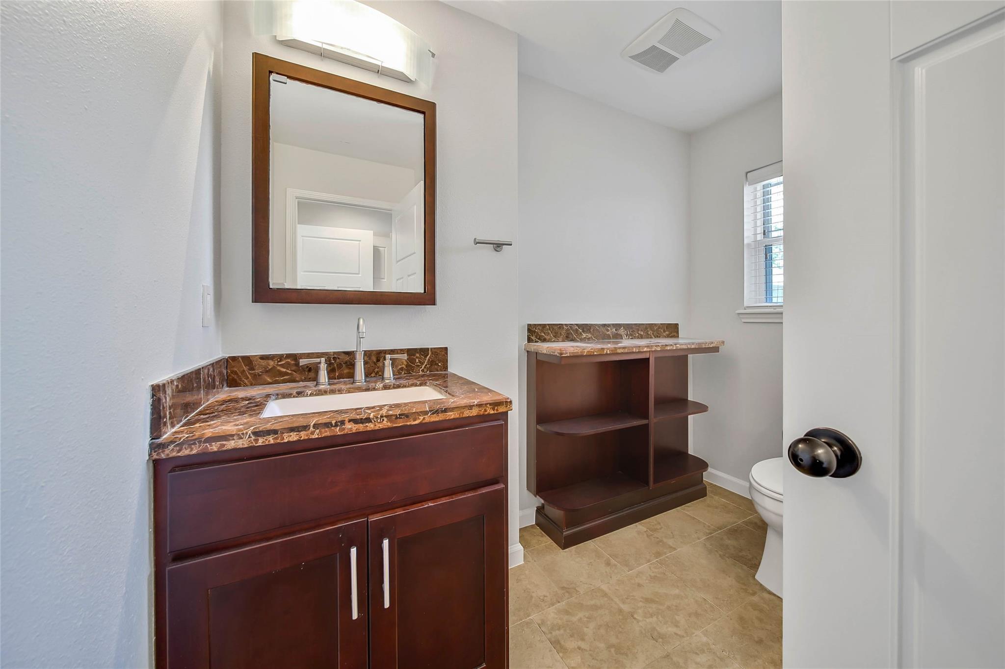 Bathroom with a dark wood vanity, brown stone-look counters, matching shelves, and a framed mirror.