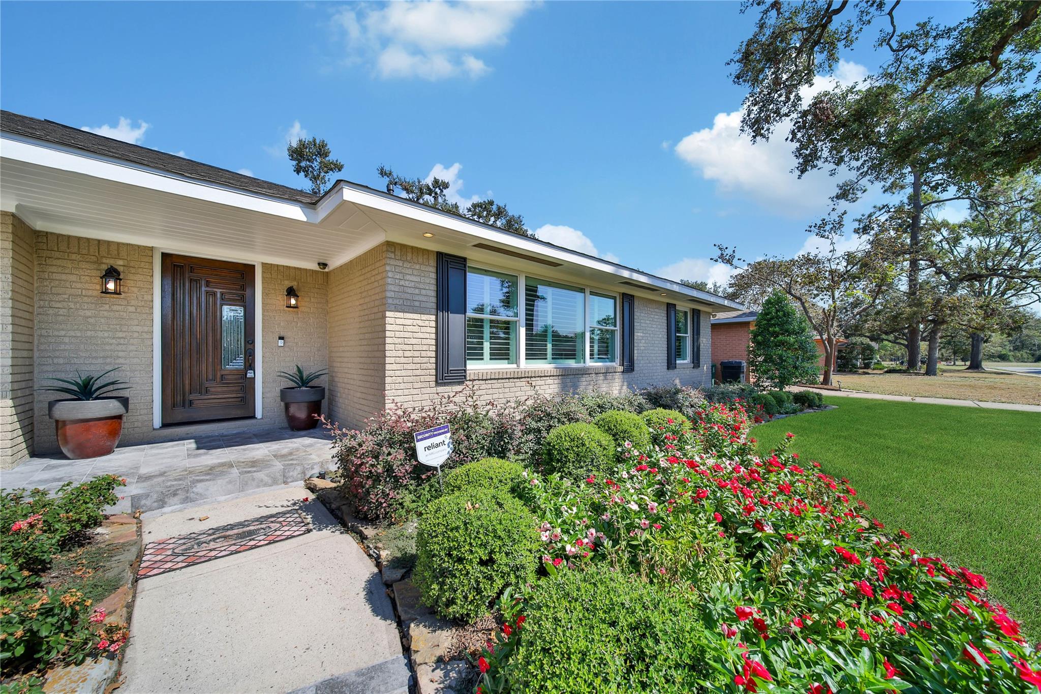 Single-story brick home with a dark wood entry, flower-filled garden beds, and a green lawn.