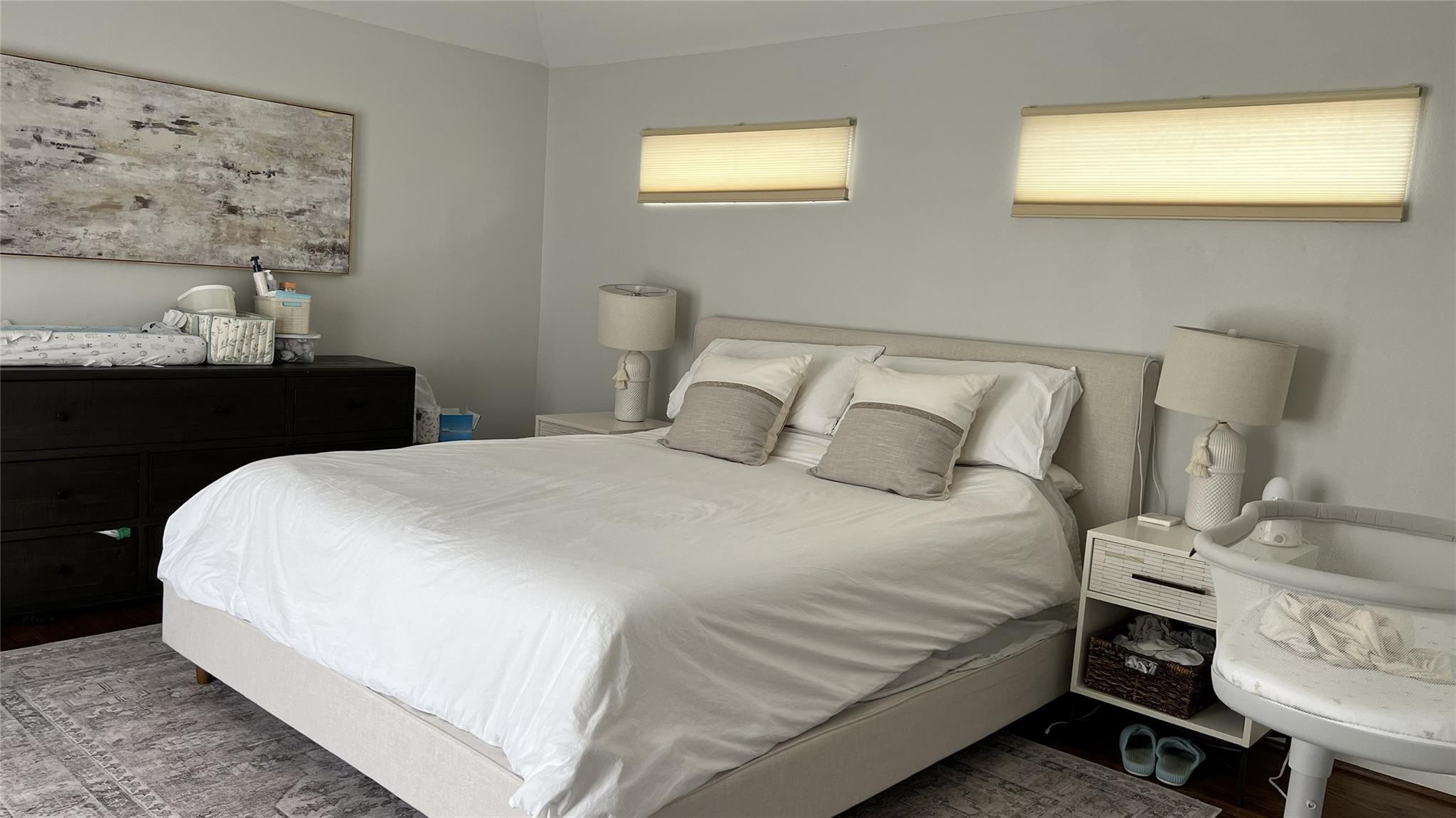Bedroom with a white-covered bed, narrow windows, dark dresser, and baby bassinet.