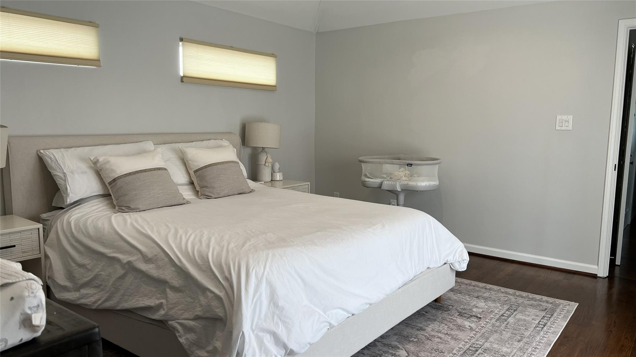 Furnished bedroom with a white bed, baby bassinet, high windows, and dark hardwood floors.