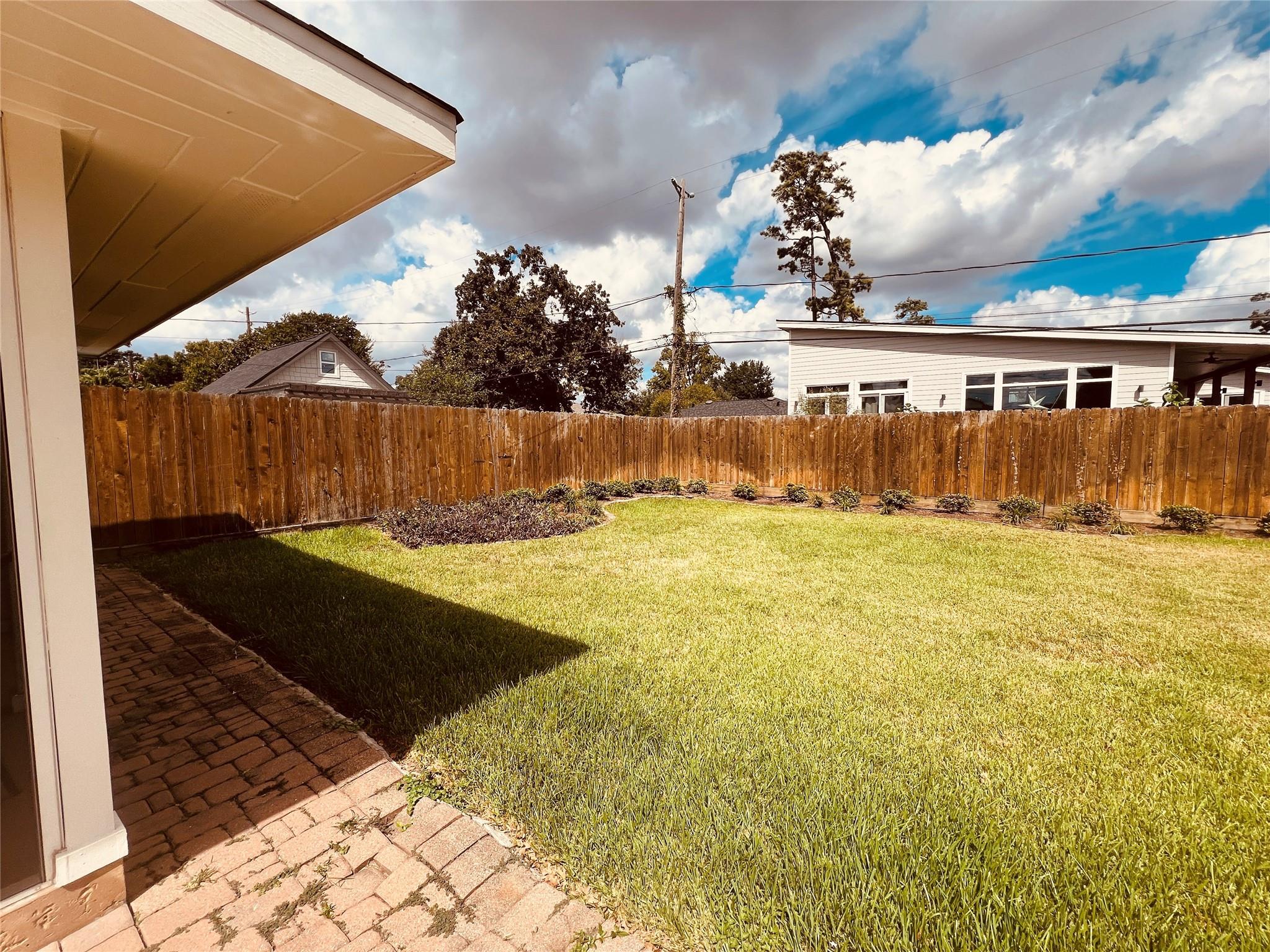 Fenced backyard with a sunny lawn, brick paver patio, and planting bed along the wooden fence.