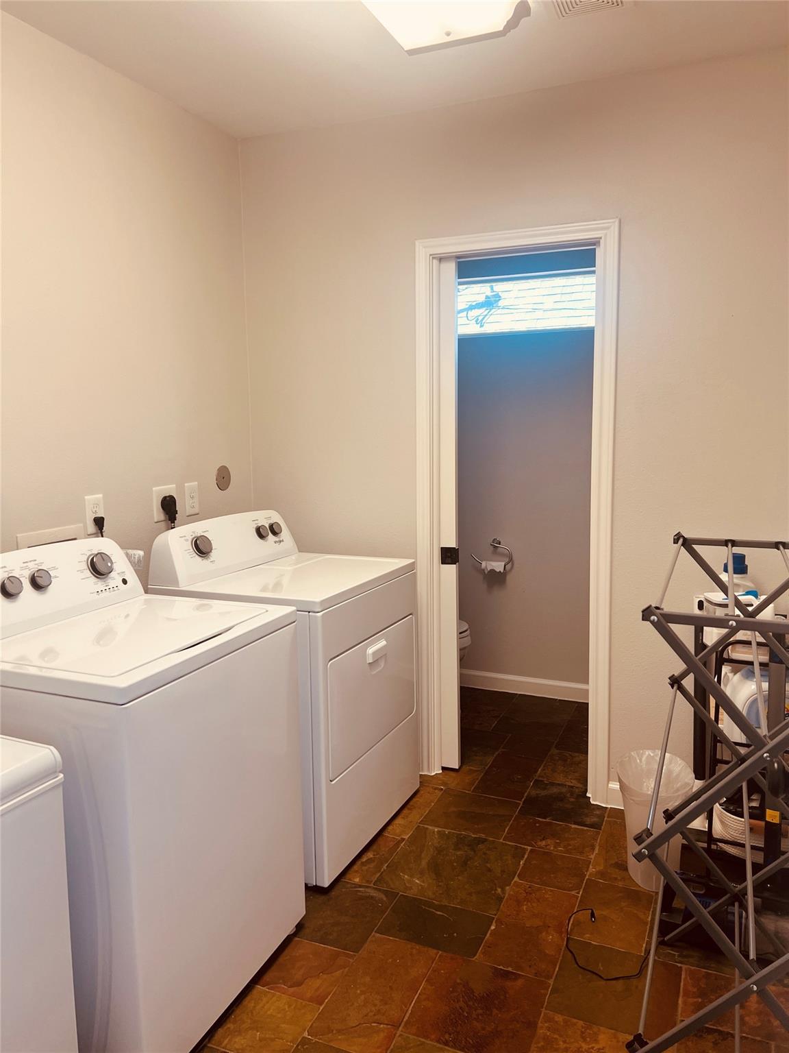 Laundry room with white washer and dryer, brown tile floor, and doorway to a bathroom.