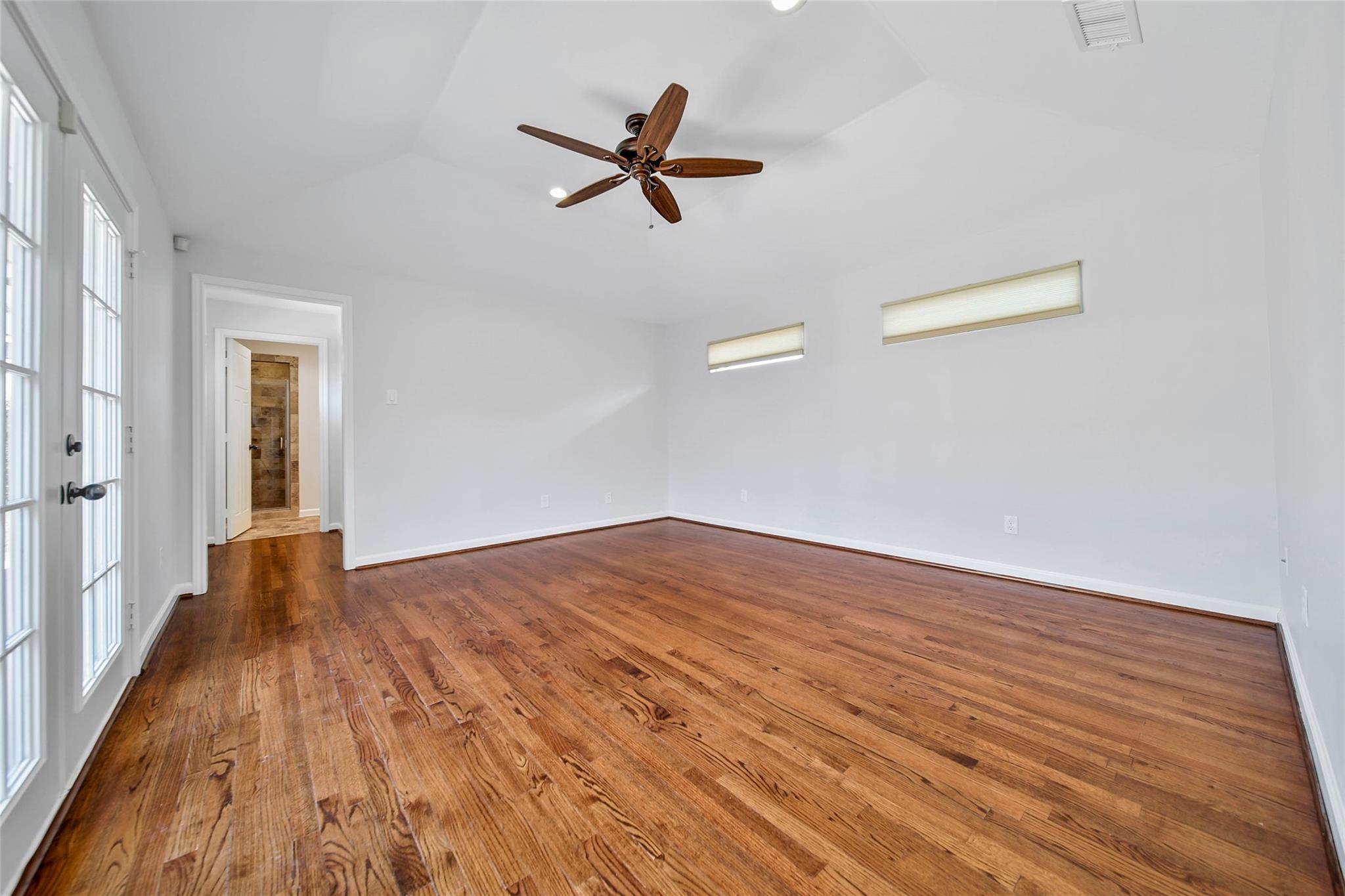 Empty room with hardwood floors, a wood-blade ceiling fan, glass-paneled doors, and narrow high windows.