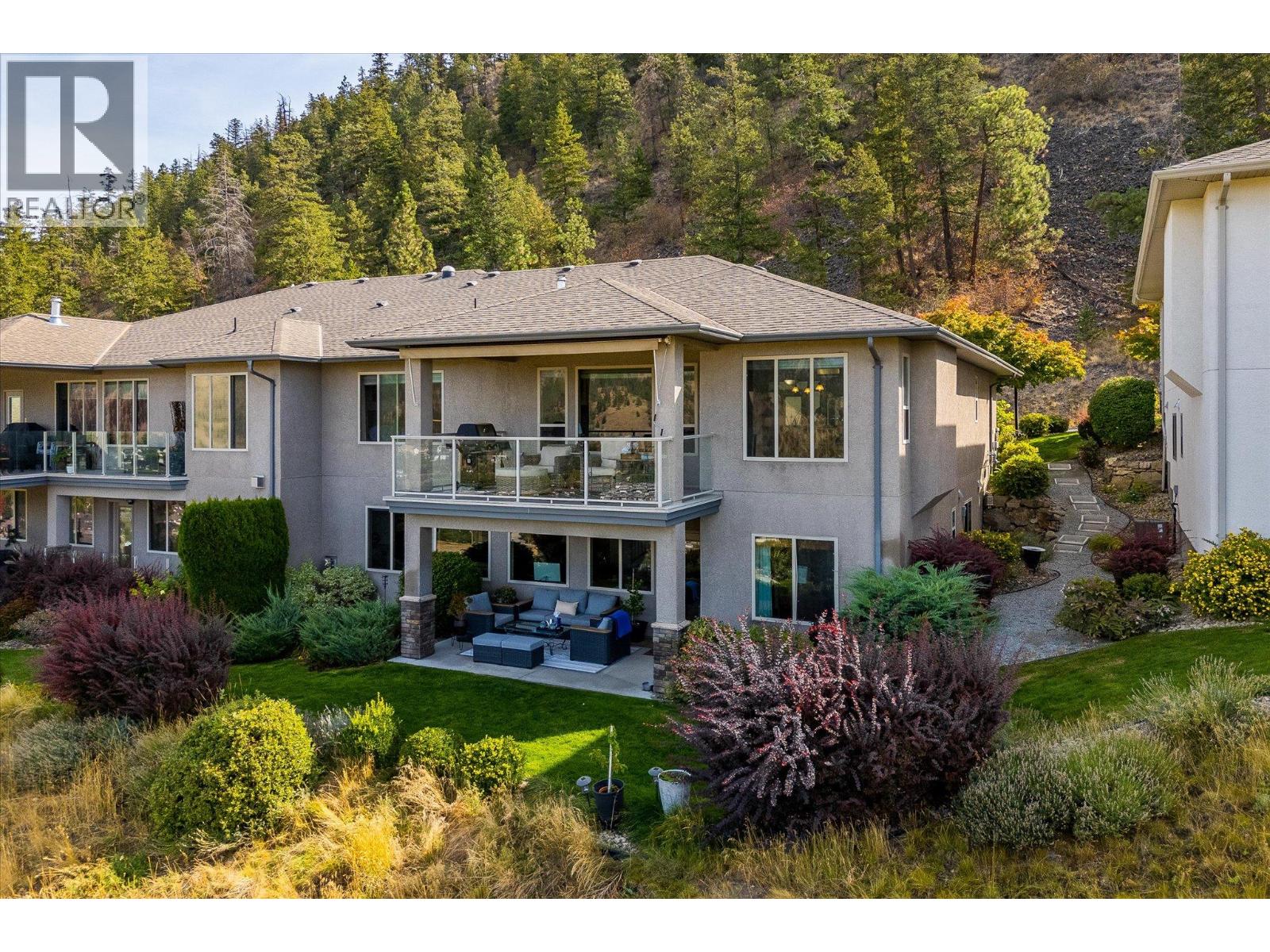 Multi-unit residence with glass-railed balconies and landscaped gardens below a wooded hillside.