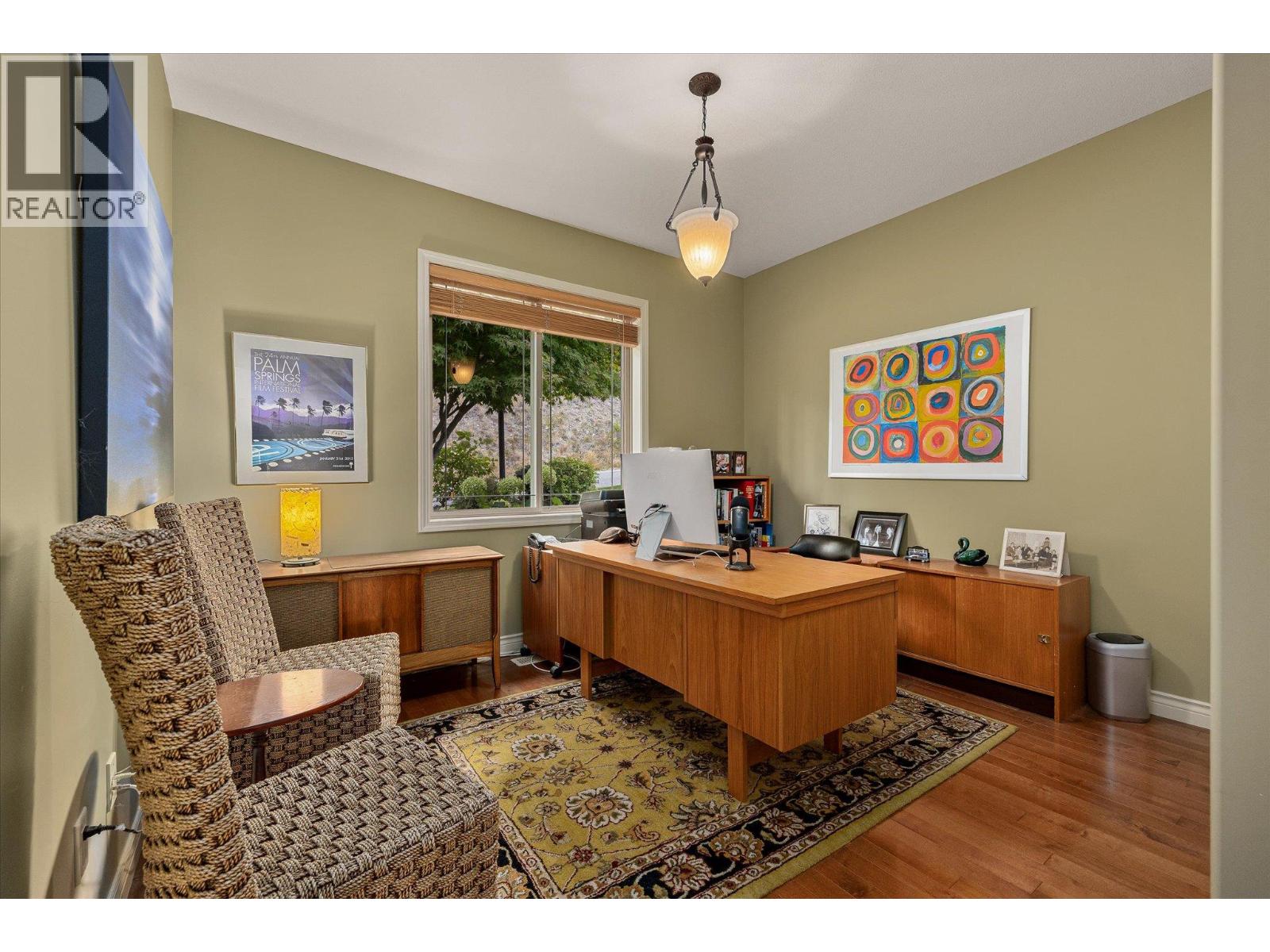 Furnished home office with a wooden desk, woven chairs, patterned rug, and window overlooking greenery.
