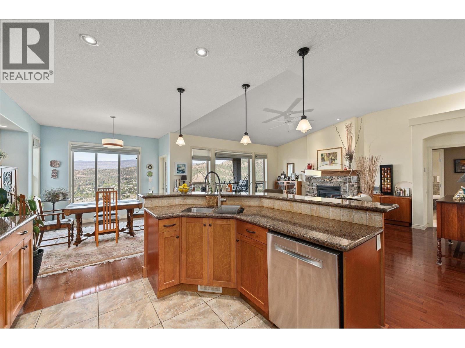 Open-plan kitchen with granite counters and wood cabinetry, dining area, stone fireplace, and wooded valley views.