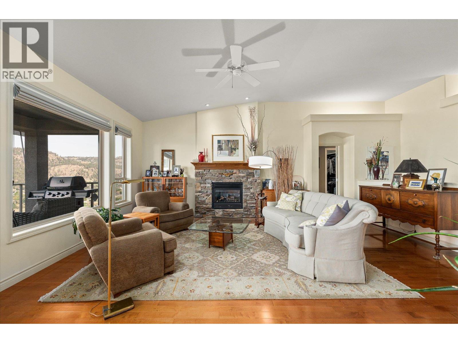 Furnished living room with stone fireplace, ceiling fan, and windows overlooking wooded hills.
