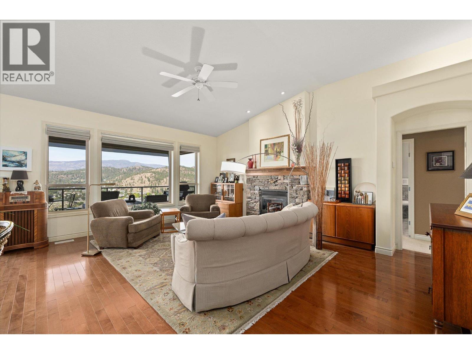 Living room with a stone fireplace, hardwood floors, and wide windows overlooking wooded hills.