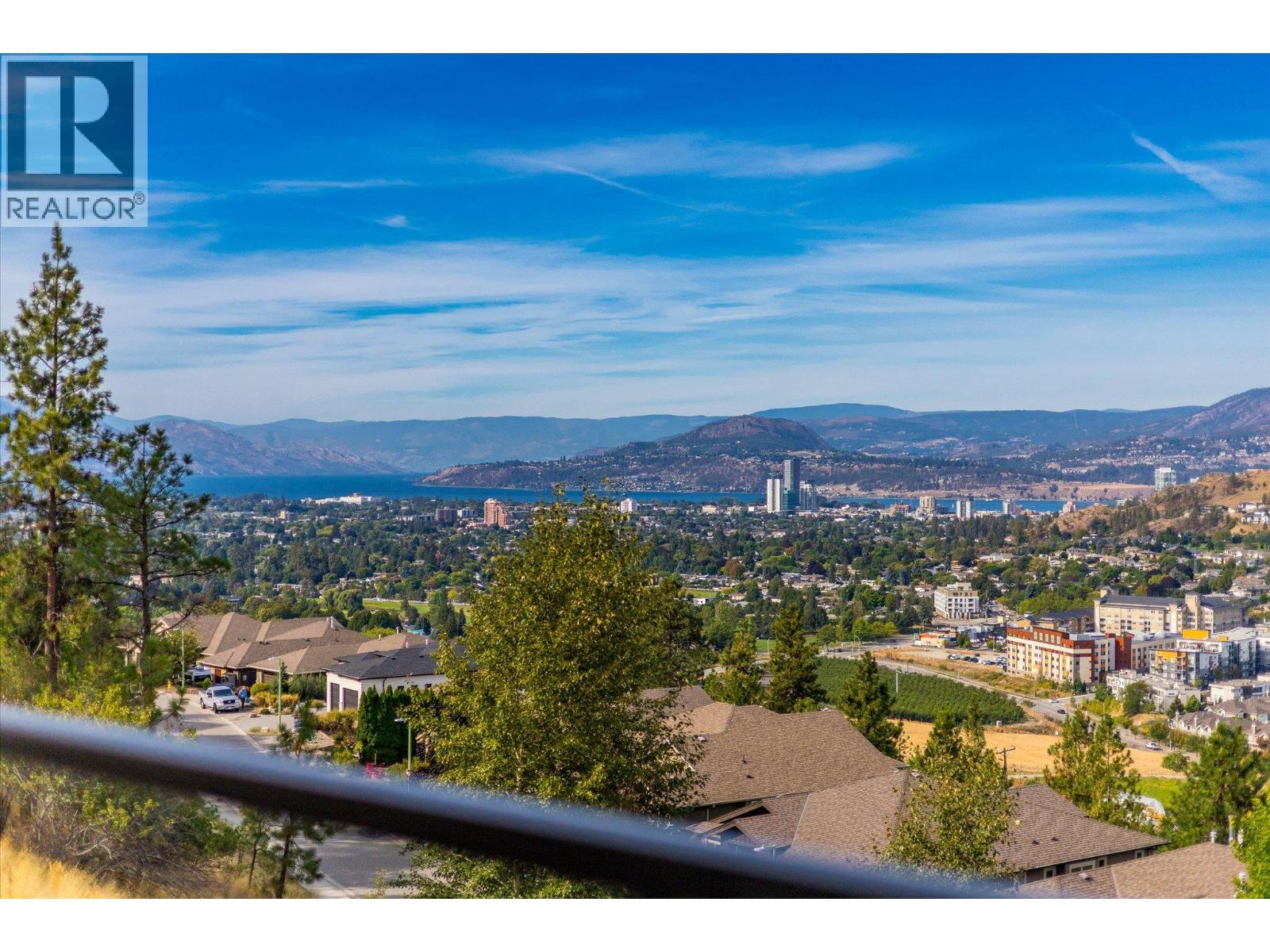 Panoramic view of Kelowna and Okanagan Lake, framed by rooftops, trees, and distant mountains.