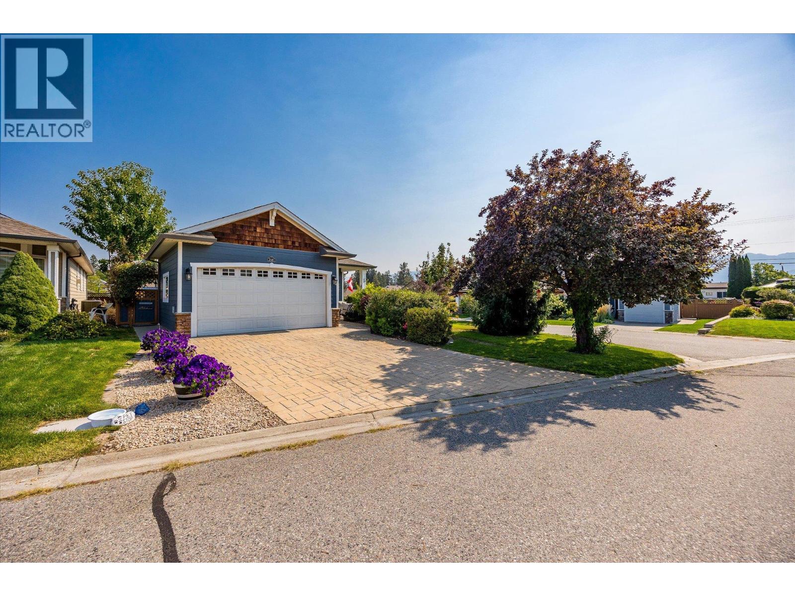 Blue-sided home with a double garage, paved driveway, purple flowers, and mature trees.