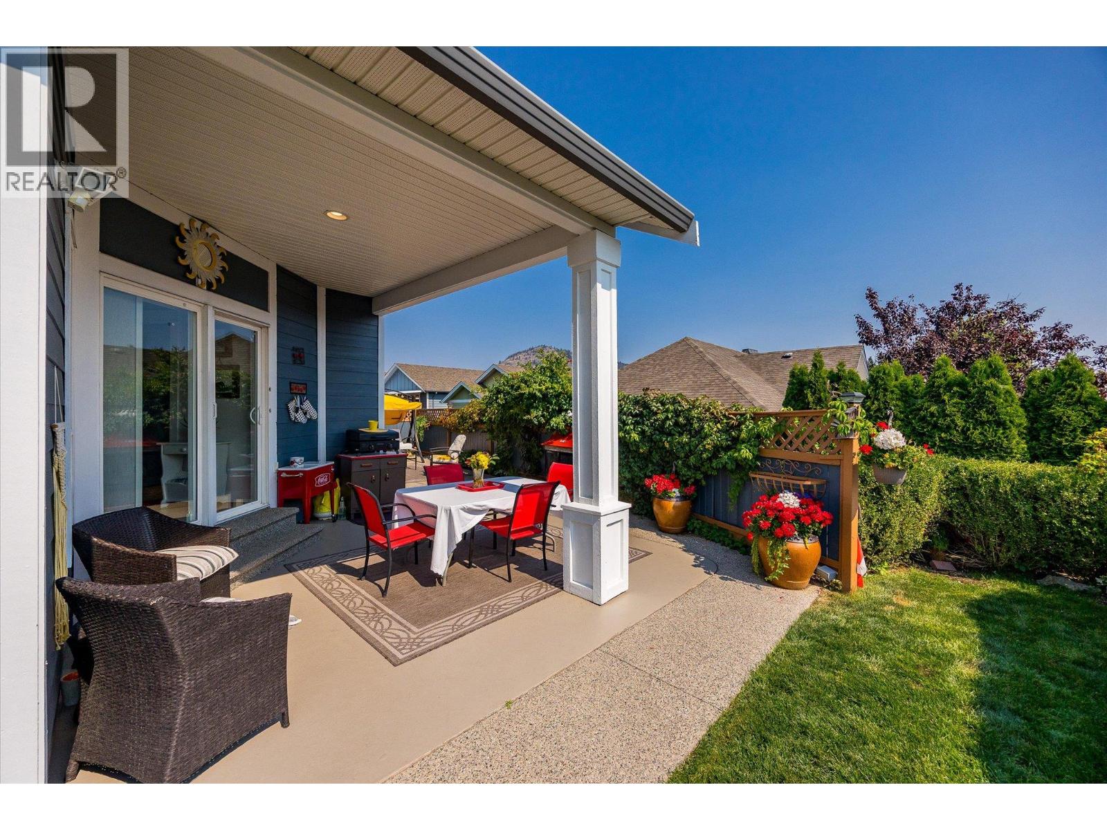 Covered backyard patio with outdoor dining set, wicker chairs, flower planters, and lawn.