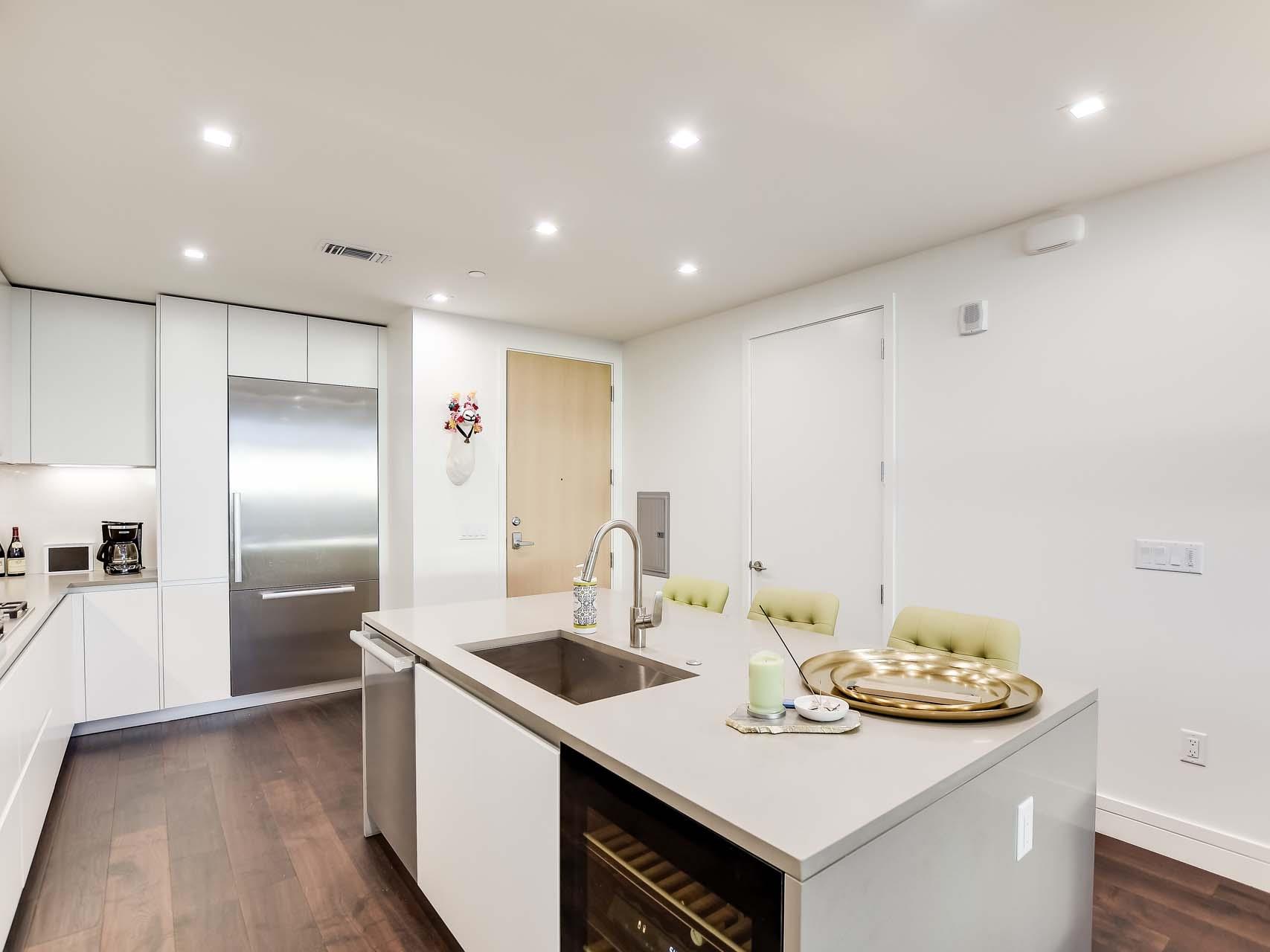Modern white kitchen with a large island, stainless-steel refrigerator, and green bar stools.