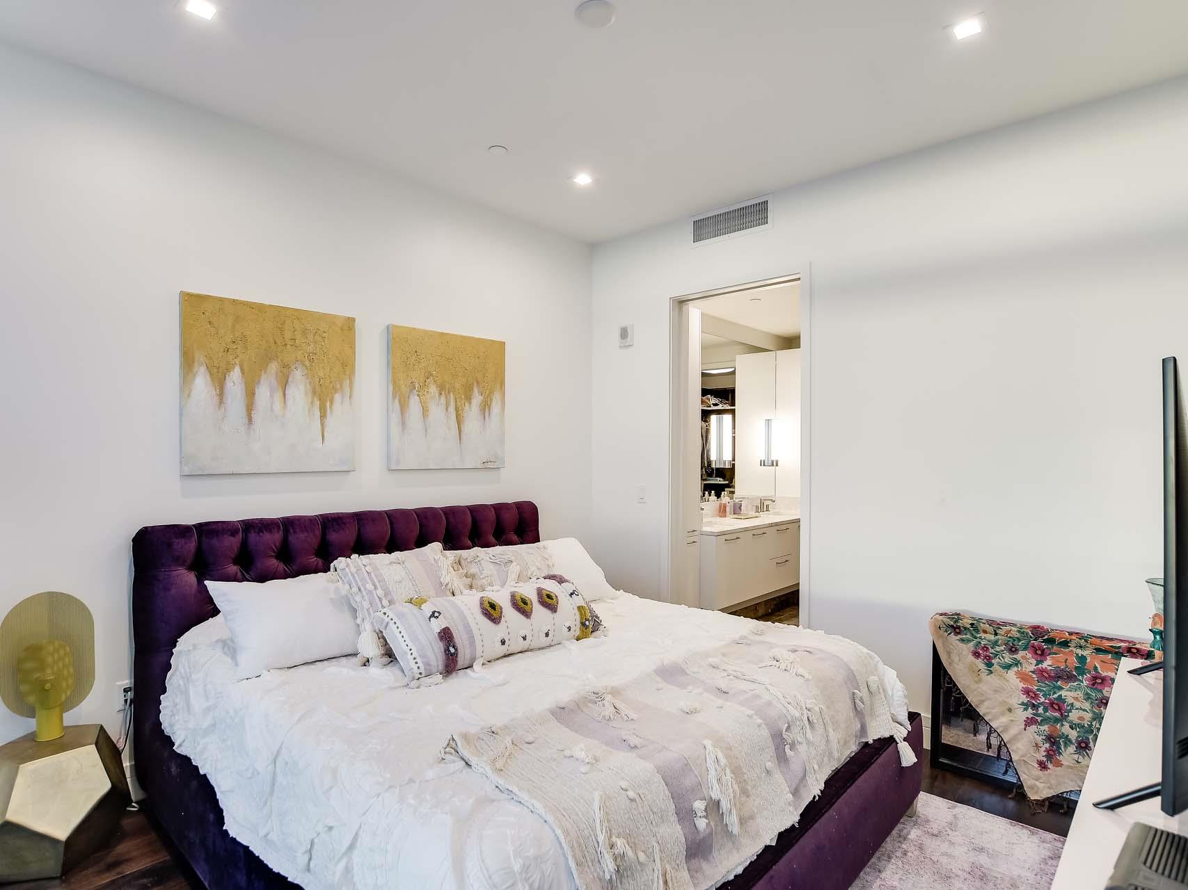 Bedroom with a white bed, purple tufted headboard, abstract wall art, and adjoining bathroom vanity.