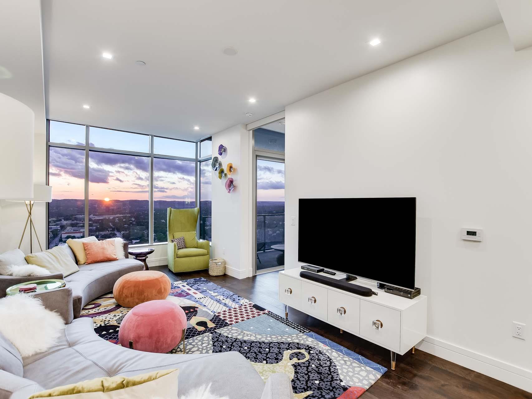 High-rise living room with corner windows overlooking the city at sunset, colorful rug, and white sectional sofas.