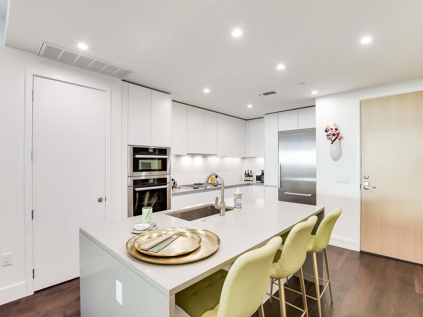 Modern white kitchen with stainless steel appliances, a large island, and pale yellow bar stools.