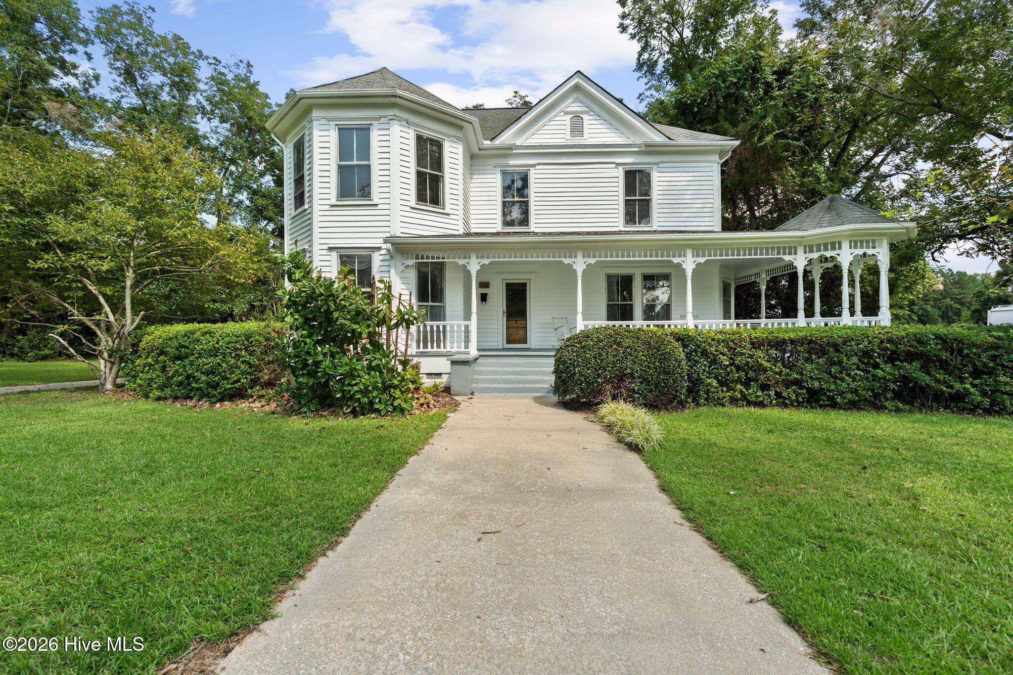 White two-story house with a wraparound porch, bay window, and walkway through a leafy lawn.