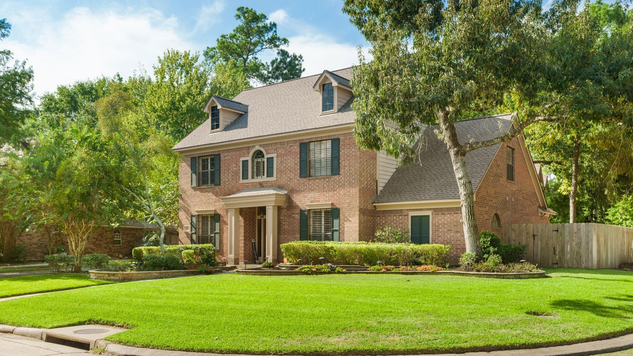 Two-story brick home with dormer windows, mature trees, trimmed hedges, and a broad front lawn.