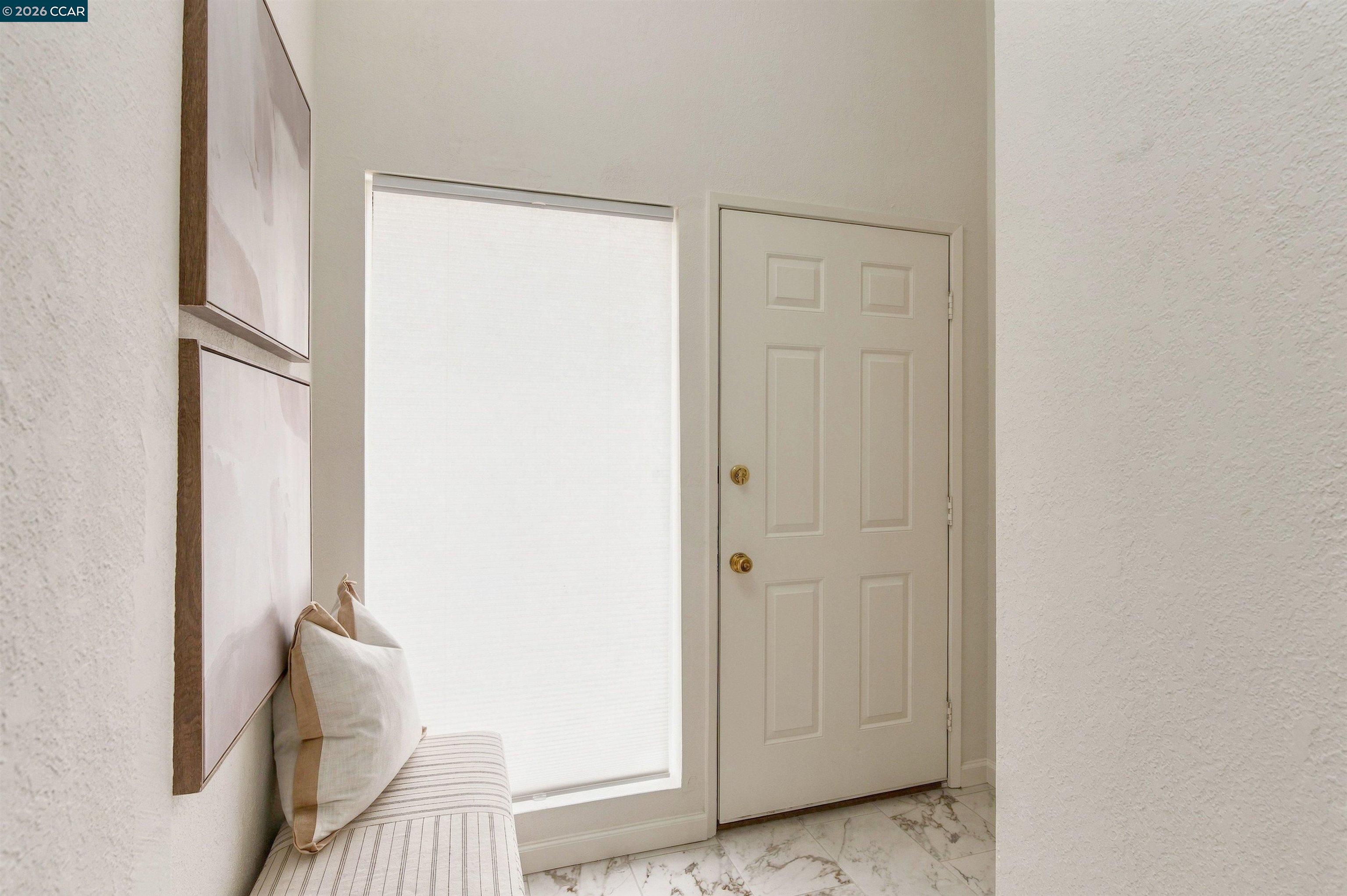 Bright entryway with a white paneled door, marble-look tile, shaded window, and upholstered bench.
