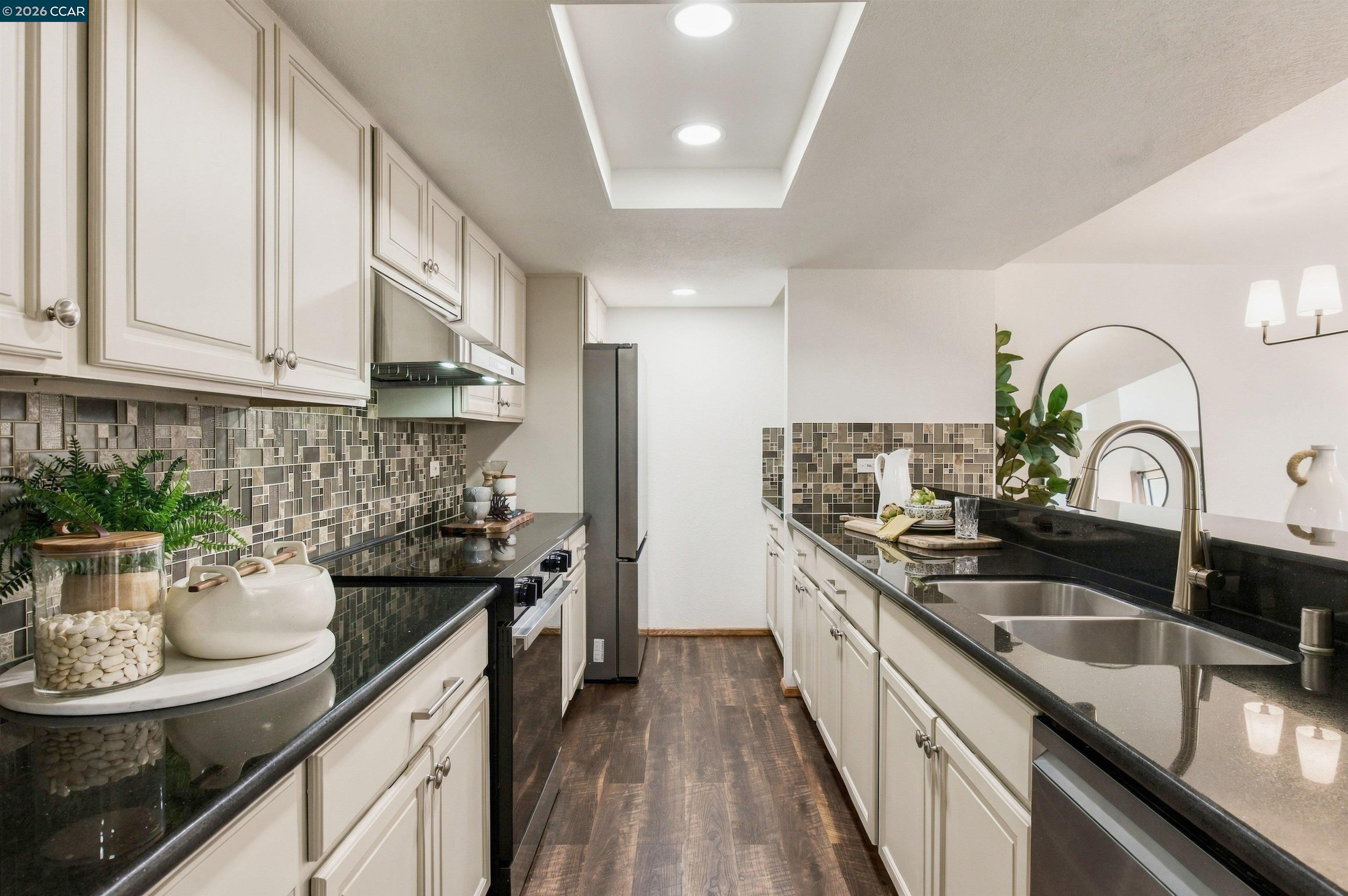 Staged galley kitchen with white cabinets, dark counters, mosaic tile backsplash, and stainless steel appliances.