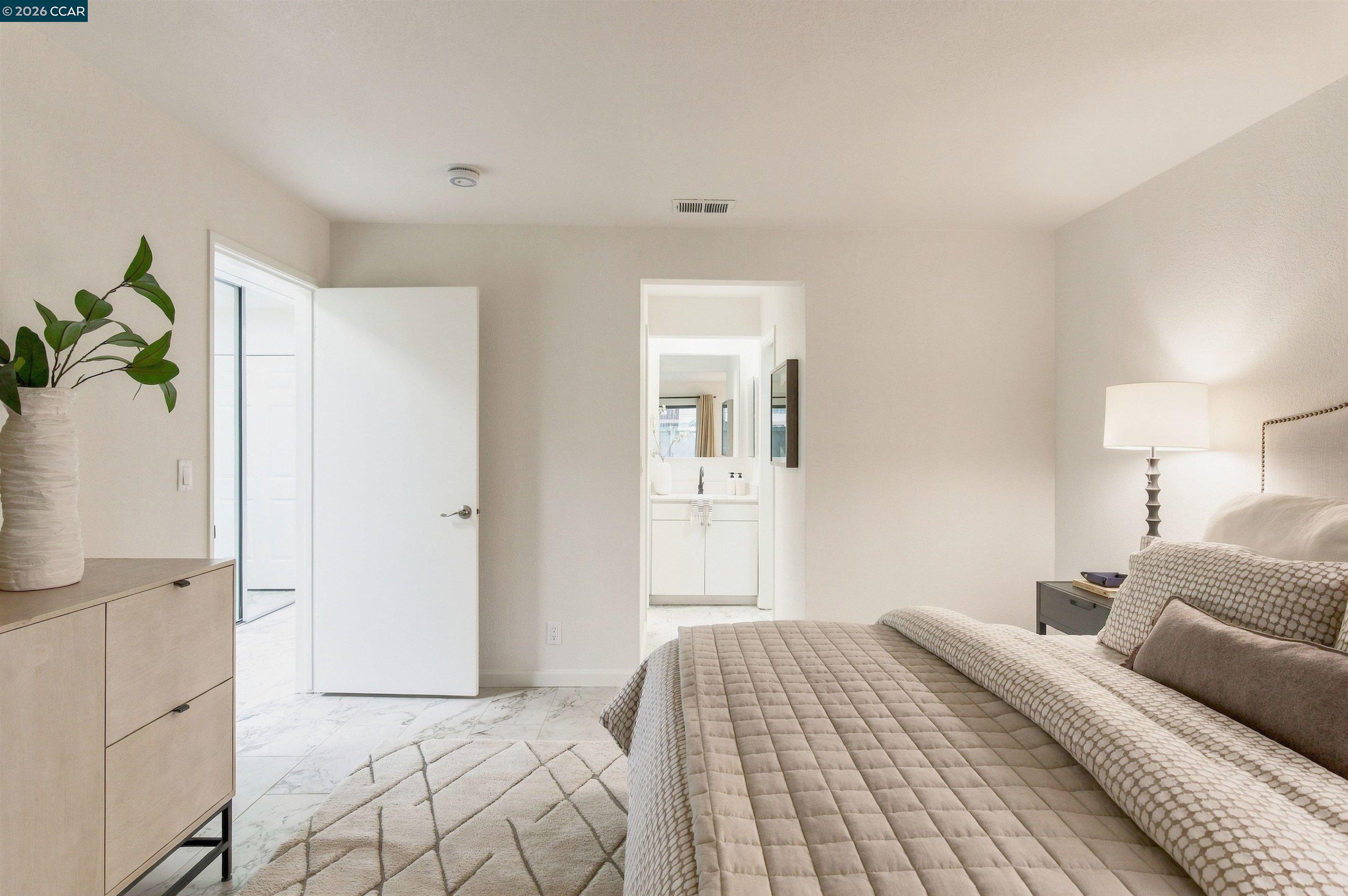 Staged bedroom with a quilted bed, light wood dresser, and open doorway to an en-suite bathroom.
