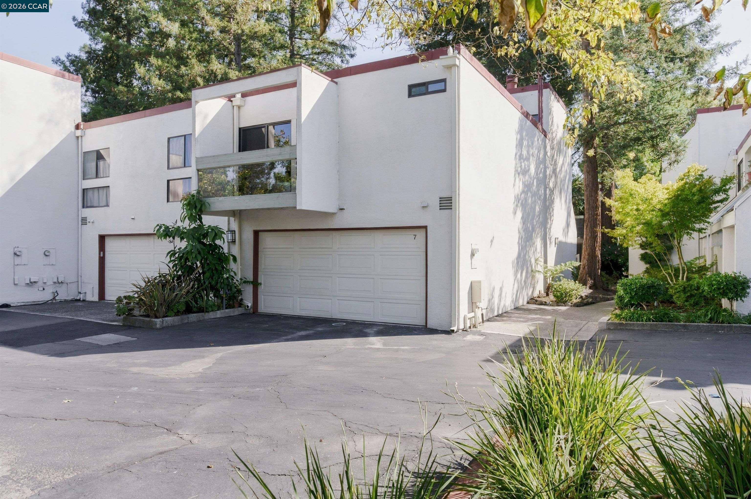 Exterior of unit 7 in a white multi-unit building, with a garage, balcony, and mature trees.