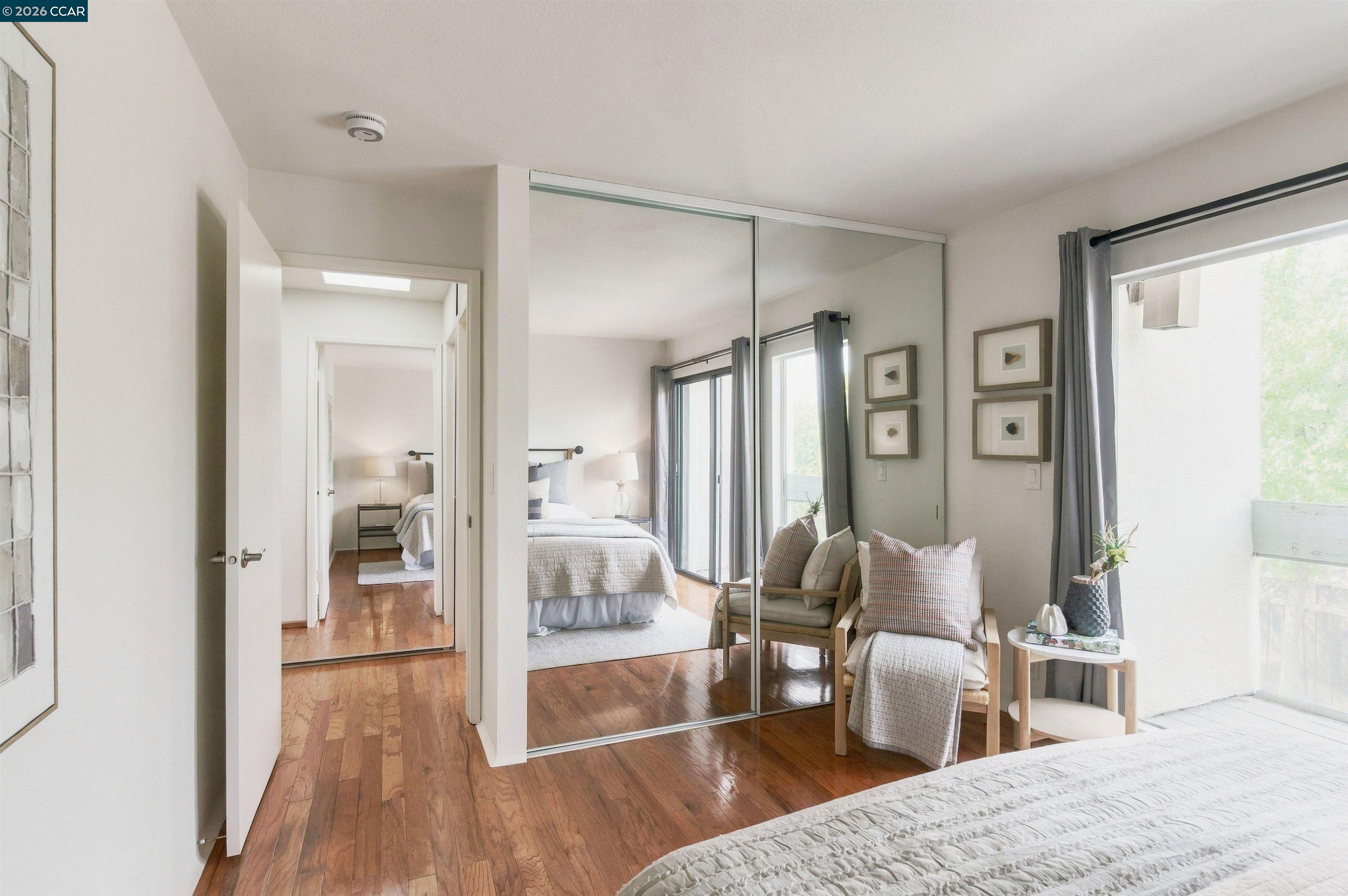 Bedroom with mirrored closet doors, hardwood floors, and a sitting area beside a large window.