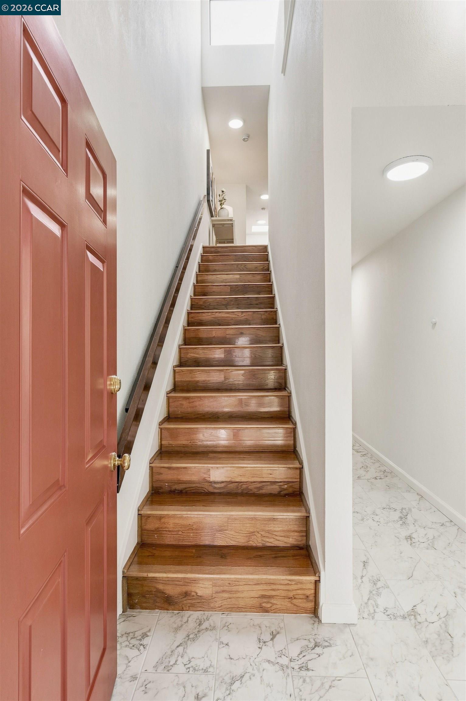 Wooden staircase with a dark handrail, beside an open reddish-brown door and marble-look tiled entryway.