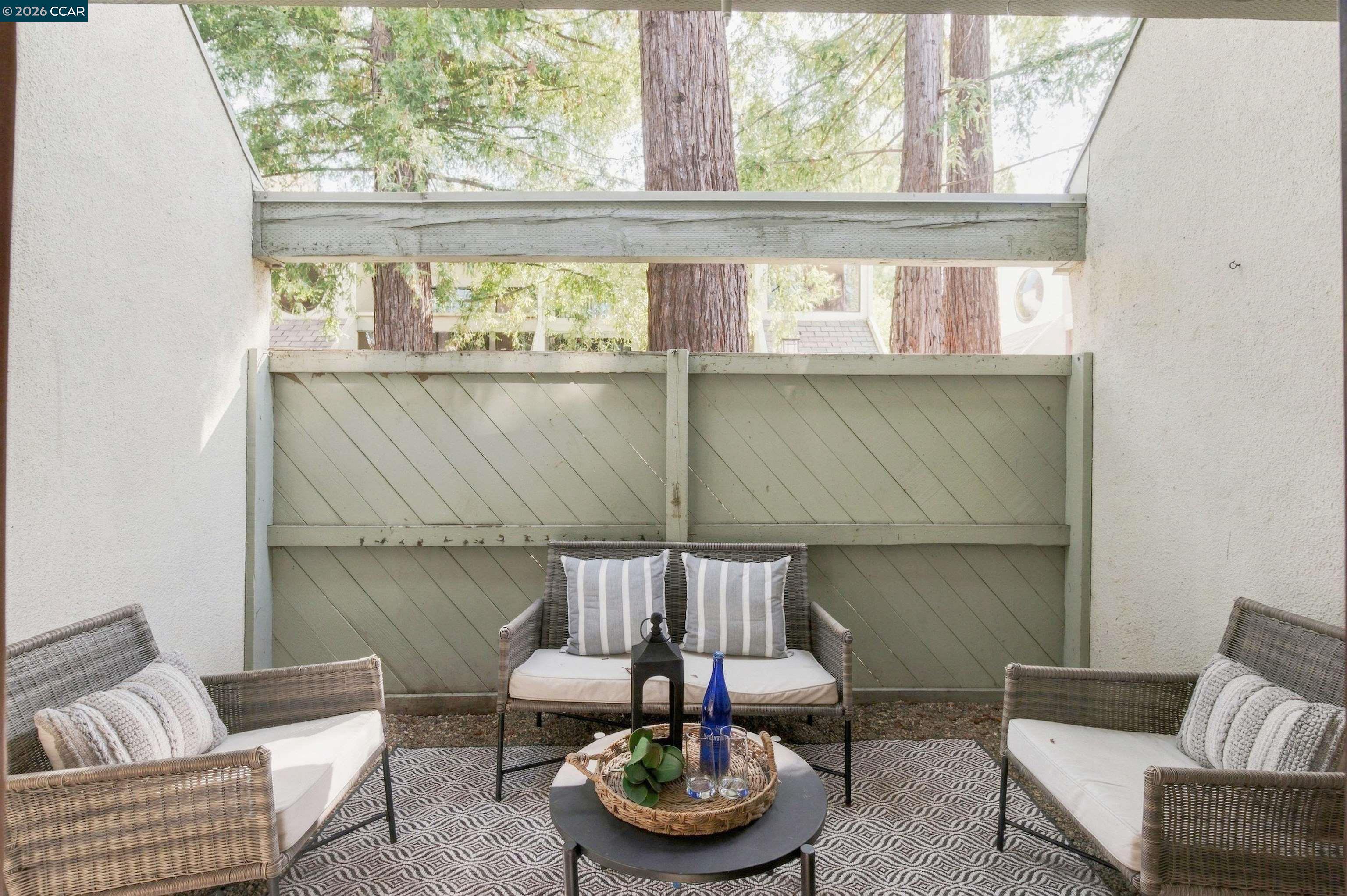 Patio with wicker seating, a patterned rug, and a green privacy fence beneath tall trees.
