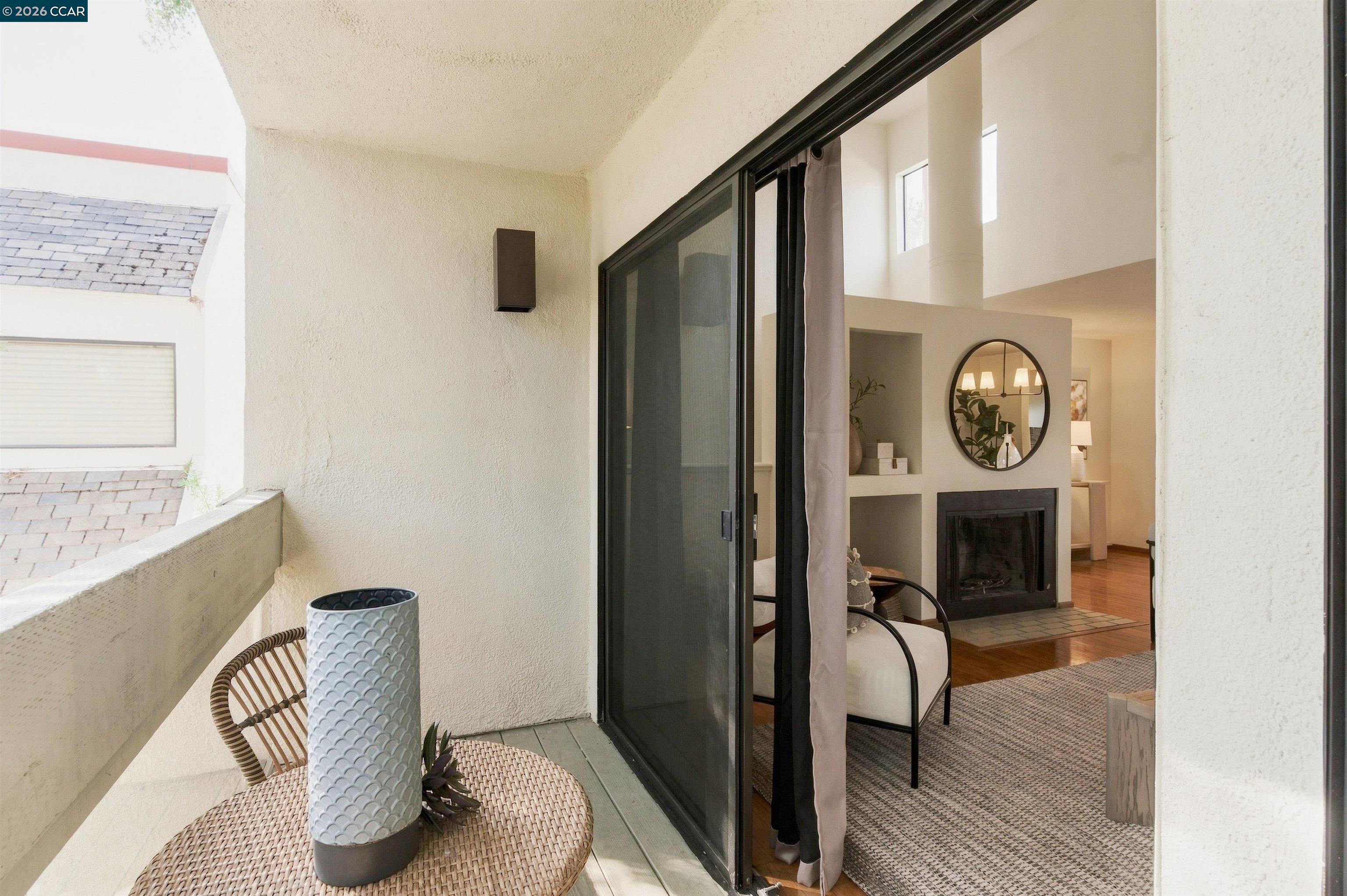 Balcony with woven furniture and planter, opening onto a staged living room with a fireplace.