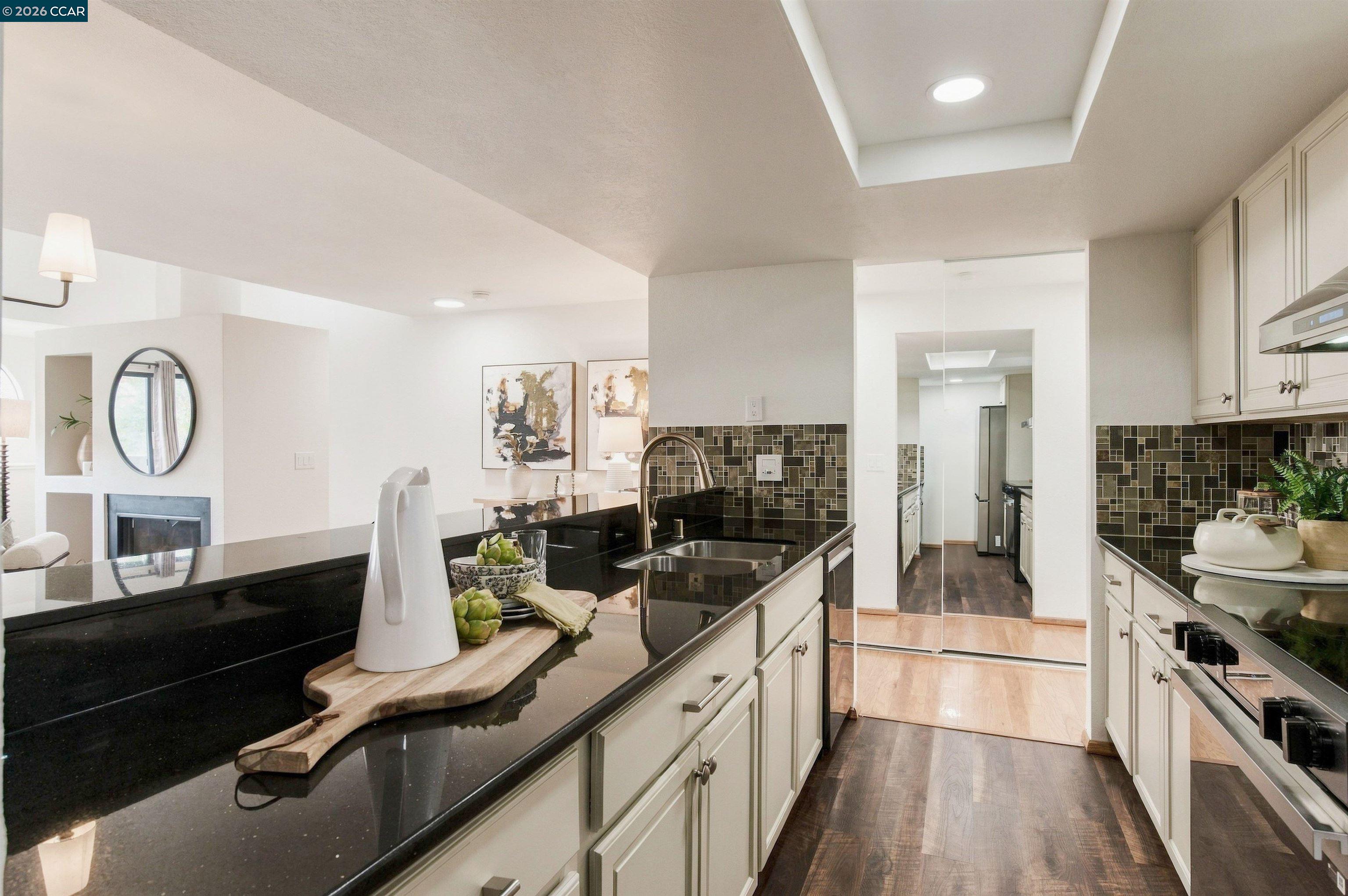 Staged kitchen with glossy black counters, mosaic tile backsplash, and a view toward the living area.