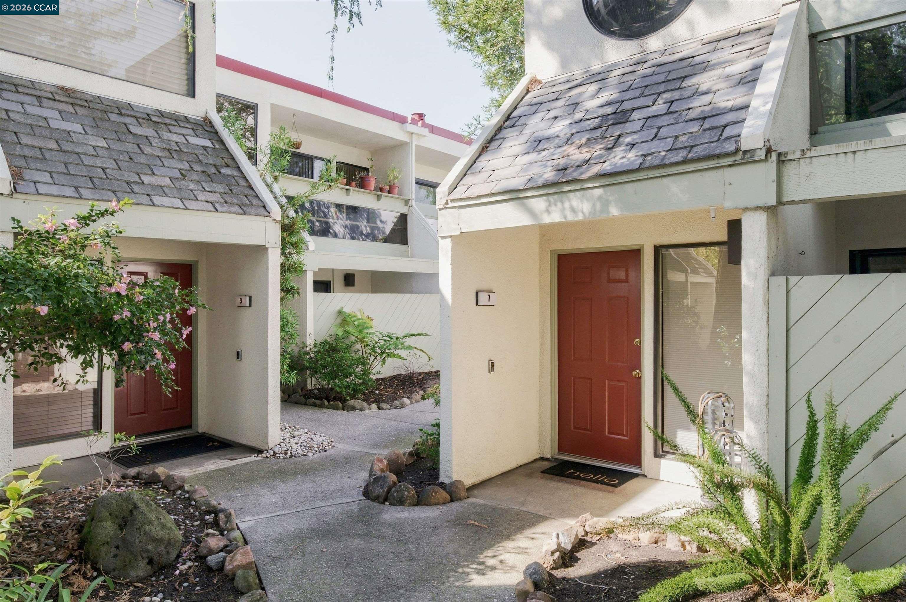 Unit 7’s red front door and shingled roof beside a landscaped walkway.