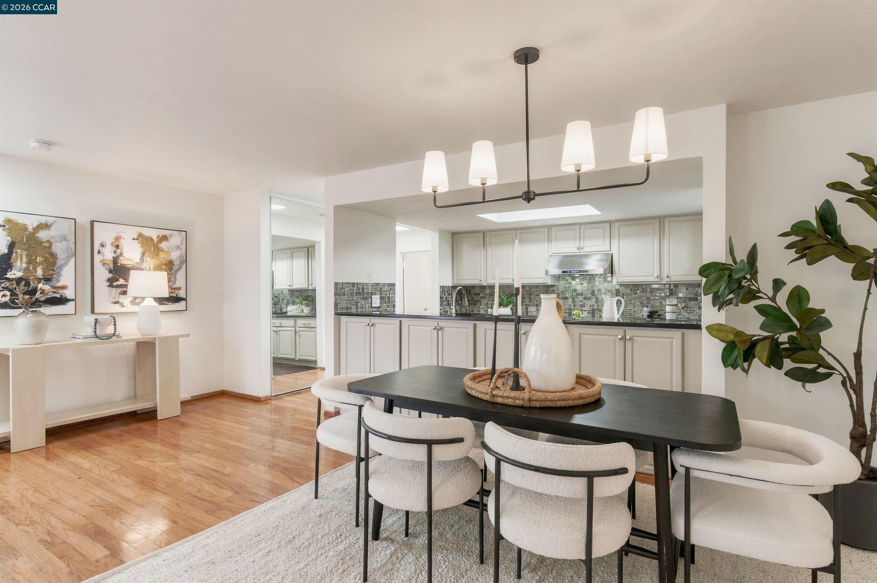 Dining area with a dark table and upholstered chairs, opening to a white kitchen with a mosaic backsplash.