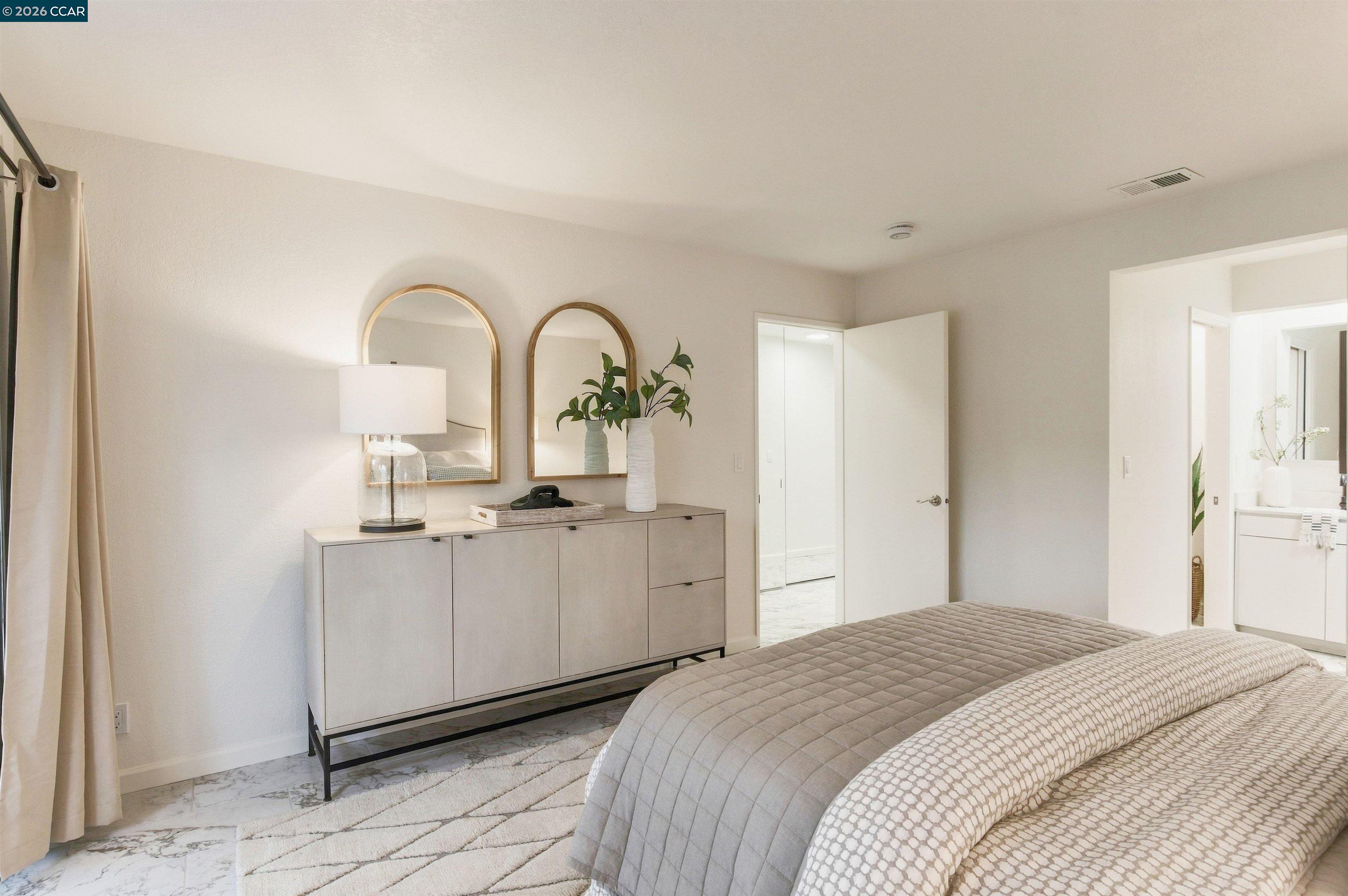 Staged bedroom with a patterned bed, arched mirrors above a dresser, and an open doorway to a bright bathroom.