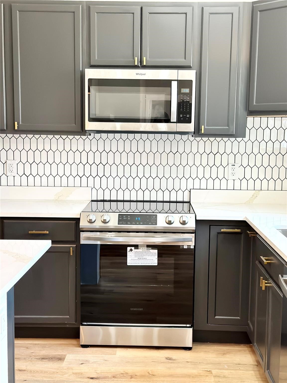 Kitchen with dark gray cabinets, geometric tile backsplash, white counters, and stainless steel appliances.