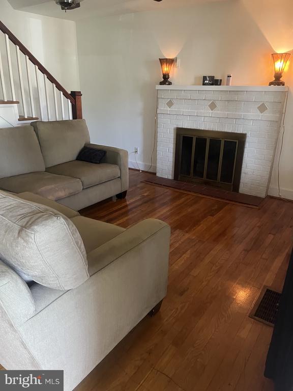 Furnished living room with white brick fireplace, hardwood floors, and staircase.