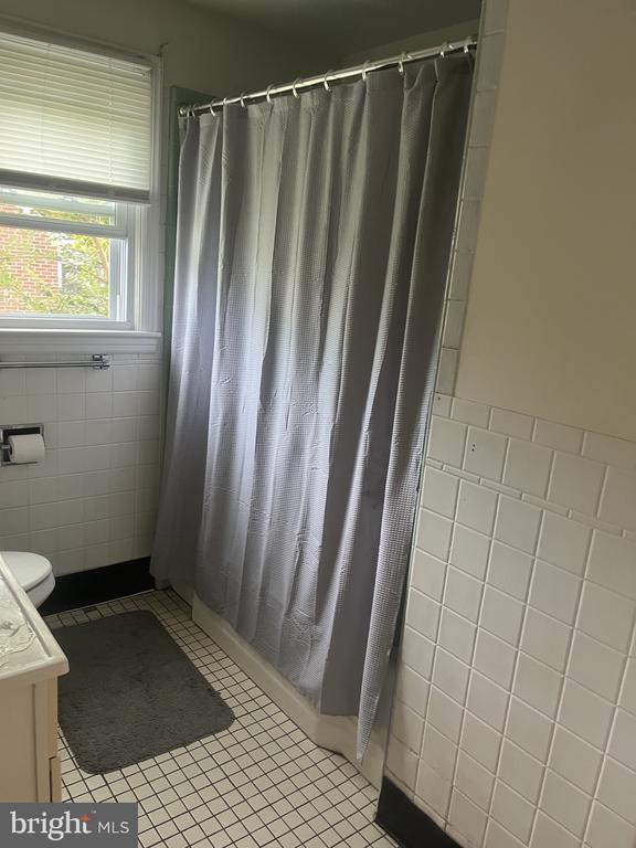 Bathroom with white tile, a gray shower curtain, and a window with blinds.