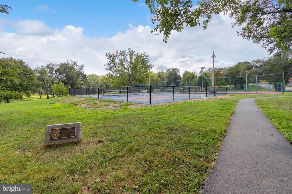 Outdoor tennis court in a grassy park, seen from a paved path with a stone marker nearby.
