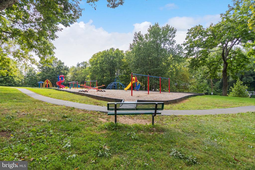 Neighborhood playground with colorful slides and swings, a bench, and a paved path under mature trees.